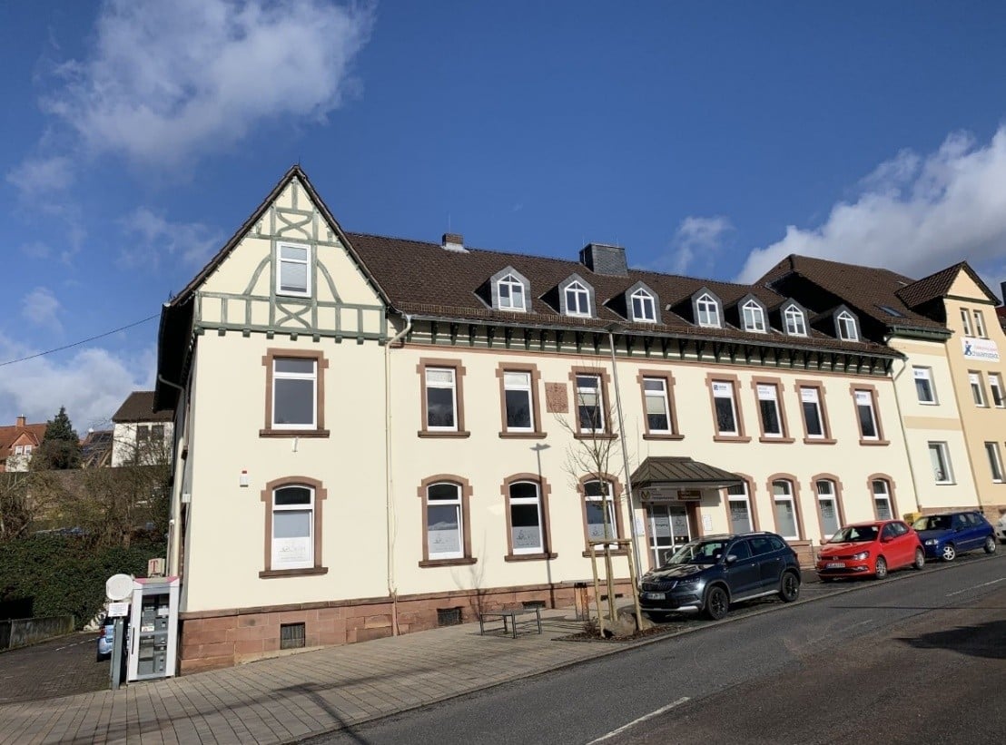 Auf dem Bild ist ein sehr großes dreigeschossiges Bürogebäude zu sehen. Die Fassade ist hell gestrichen. Die Fenster in allen Geschossen sind symmetrisch über die ganze Front des Hauses angeordnet und farbig abgesetzt. In der Mitte des Erdgeschosses befindet sich ein überdachter Eingangsbereich mit Automatiktür. Im Dachgeschoss wurden sieben Gauben eingebaut. Das Dach ist mit dunklen Ziegensteinen eingedeckt. Im rechten Bildrand erkennt am einen modernen Anbau an das vorhandene unter Denkmalschutz stehende Gebäude. Vor dem Gebäude befinden sich drei PKW-Stellplätze, eine Sitzbank und ein Baum. Links neben der Immobilie erkennt man eine Einfahrt zum dahinter liegenden Hof, sowie eine Telefonzelle. Das Haus ist nach Süden ausgerichtet, so dass die Büroräume lichtdurchflutet sind.