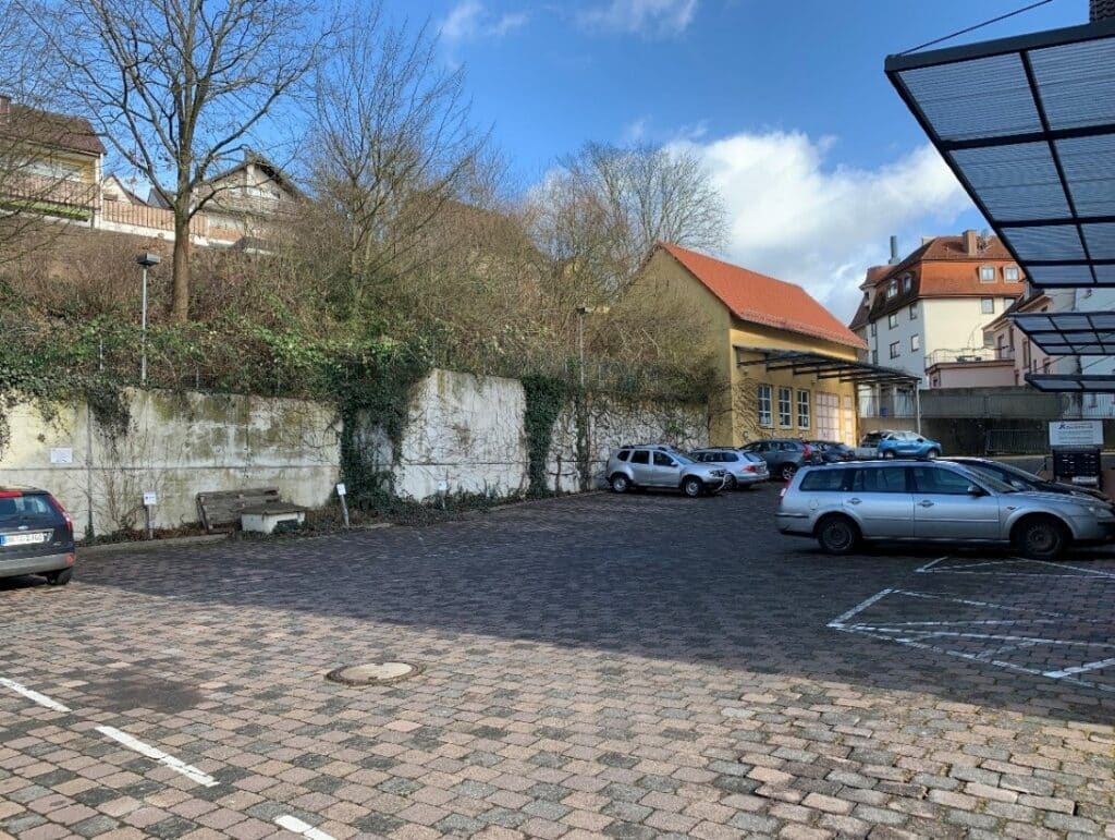 Auf dem Foto ist ein sehr großzügig gepflasterter Hinterhof mit einigen geparkten Autos zu sehen. Begrenzt wird der Hof durch eine hohe Mauer im rückwärtigen Bereich, da das Gelände stark ansteigt. Von dem Gebäude sich nur drei Vordächer in Teilen zu sehen. Im hinteren Bereich erkennt man noch ein kleineres in gelb gestrichenes Gebäude mit Vordach und drei großen Sprossenfenstern. Dieses wird zurzeit als Lagerfläche genutzt.