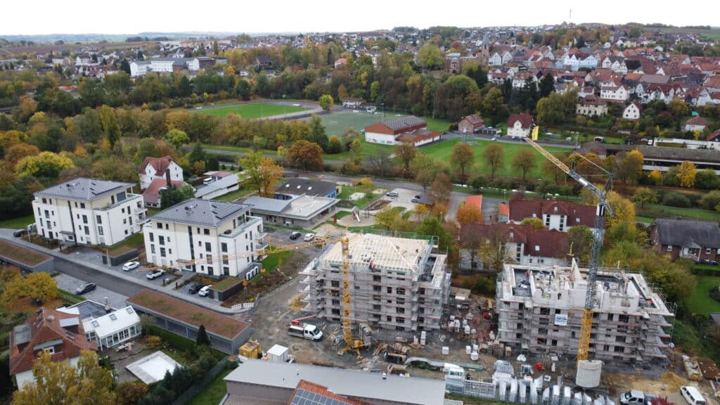 Das Bild zeigt eine Luftaufnahme eines Wohngebiets, in dem eine Baustelle im Vordergrund dominiert. Im Vordergrund befinden sich zwei im Bau befindliche mehrstöckige Gebäude. Diese sind von Gerüsten umgeben und werden von einem gelben Kran bedient. Auf den Dächern der Gebäude sind Arbeiter zu erkennen. Die Bauarbeiten sind in vollem Gange. Neben den im Bau befindlichen Gebäuden befinden sich bereits fertige Wohngebäude mit modernen Designs und Flachdächern. Einige Häuser haben Solarpanels auf ihren Dächern. Im Hintergrund erstreckt sich ein weitläufiges Wohngebiet mit einer Mischung aus traditionellen Häusern mit roten Dächern und neueren Gebäuden. Ein Fußballplatz und ein Sportplatz sind ebenfalls zu sehen, umgeben von Bäumen, die sich im Herbst verfärben.