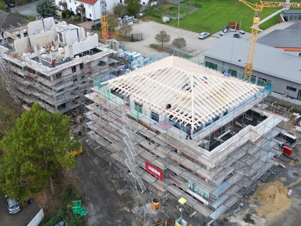 Das Bild zeigt eine Baustelle aus der Vogelperspektive. Im Zentrum stehen zwei Gebäude, die sich im Bau befinden. Das Hauptgebäude, das sich im Bau befindet, ist ein mehrstöckiges Gebäude mit einem Holzrahmen, der gerade für das Dach gebaut wird. Gerüste umgeben das Gebäude und ermöglichen es Arbeitern, verschiedene Bereiche zu erreichen. Arbeiter sind auf dem Dach und an den Gerüsten zu sehen. Links vom Hauptgebäude befindet sich ein weiteres Gebäude, das ebenfalls im Bau ist, aber in einem früheren Stadium. Der Bau von Wänden und anderen Strukturen ist erkennbar. Ein gelber Kran steht auf der rechten Seite des Bildes und wird vermutlich zum Heben von Baumaterialien verwendet. Die Farben im Bild sind überwiegend gedämpft, mit Grautönen für die Gebäude und Gerüste. Das Holz der Dachkonstruktion ist hell, während der grüne Rasen und einige Bäume Farbtupfer hinzufügen. Der Himmel ist bedeckt und leicht getrübt.