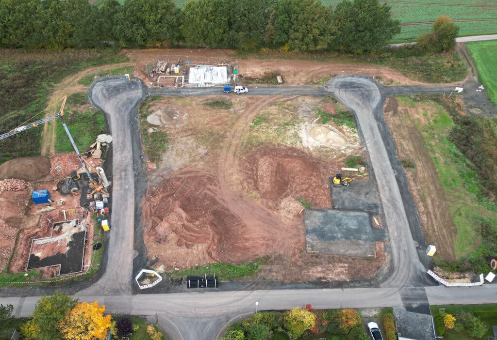 Das Bild zeigt eine Baustelle aus der Vogelperspektive. Es ist eine Szene mit roter Erde, die von Baumaschinen befahren wurde. Es gibt zwei Straßen, die in die Baustelle führen, die sich im Bau befindet. Auf der linken Seite befindet sich ein Kran, der sich über die Grube erstreckt, und ein Bagger. Ein Fundament ist in der Grube zu sehen. In der Nähe der Grube stehen Baucontainer. Auf der rechten Seite der Baustelle befinden sich bereits fertige Straßenabschnitte. Ein kleiner Bagger ist zu sehen. Auf der Baustelle fahren ein weißer Transporter mit Anhänger. Im Hintergrund befinden sich grüne Felder und ein Wald. Das Wetter scheint bewölkt zu sein.
