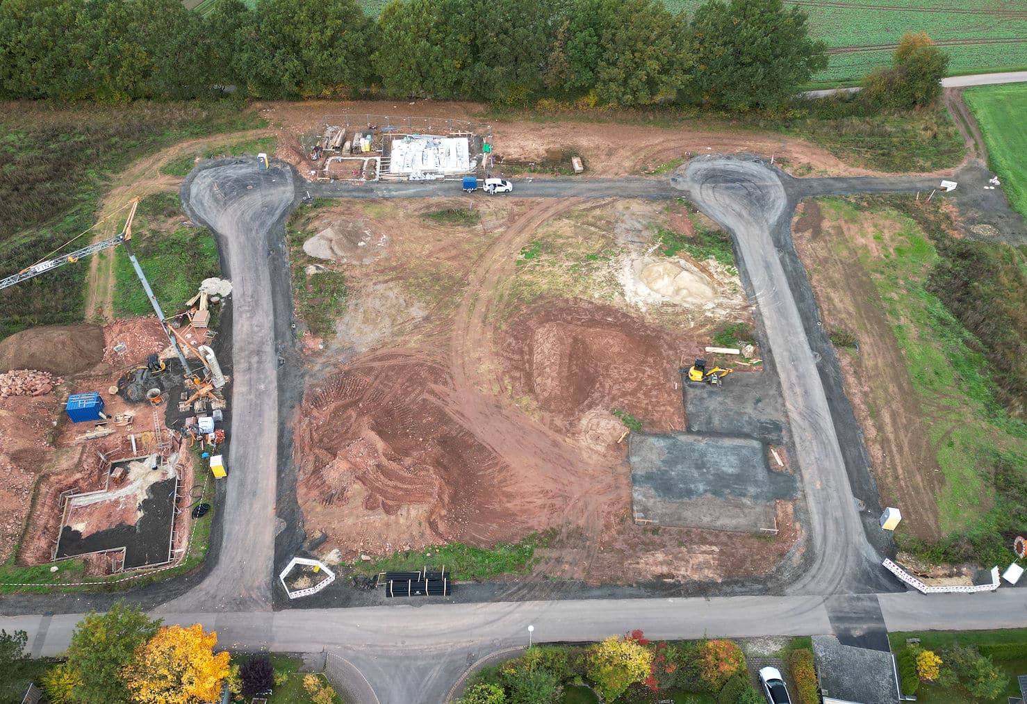 Das Bild zeigt eine Baustelle aus der Vogelperspektive. Es ist eine Szene mit roter Erde, die von Baumaschinen befahren wurde. Es gibt zwei Straßen, die in die Baustelle führen, die sich im Bau befindet. Auf der linken Seite befindet sich ein Kran, der sich über die Grube erstreckt, und ein Bagger. Ein Fundament ist in der Grube zu sehen. In der Nähe der Grube stehen Baucontainer. Auf der rechten Seite der Baustelle befinden sich bereits fertige Straßenabschnitte. Ein kleiner Bagger ist zu sehen. Auf der Baustelle fahren ein weißer Transporter mit Anhänger. Im Hintergrund befinden sich grüne Felder und ein Wald. Das Wetter scheint bewölkt zu sein.