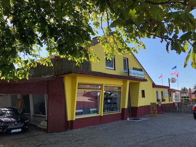 Das Bild zeigt ein Gebäude mit einem auffälligen Farbschema. Das Gebäude selbst ist in einem leuchtenden Gelbton gehalten, der durch dunkelbraune Akzente ergänzt wird, insbesondere an der Fassade und am Dach. Das Gebäude ist von einem Baum mit üppigem, grünem Laub umgeben, das einen natürlichen Rahmen für das Bild bildet. Das Sonnenlicht scheint durch die Blätter. Vor dem Gebäude befindet sich ein Parkplatz, der mit grobem Kies bedeckt ist. Ein schwarzes Auto ist am linken Rand des Bildes geparkt. Am Gebäude sind verschiedene Schilder und Dekorationen angebracht, die auf die Nutzung des Gebäudes als Gastronomiebetrieb hindeuten. Ein Schild mit der Aufschrift "Mannis Ostseestube" ist über dem Eingang angebracht. Kleine Fahnen, darunter die deutsche und eine europäische Flagge, sowie ein Schild mit einem Bierlogo, sind vor dem Gebäude gehisst. Die Komposition des Bildes ist ausgewogen, mit dem Gebäude als zentralem Fokus und dem Baumlaub, das einen natürlichen Rahmen bildet.