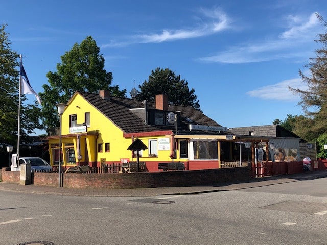 Das Bild zeigt ein zweistöckiges Gebäude, das sich an einer Straßenecke befindet. Das Gebäude ist in einem leuchtenden Gelb gestrichen, mit dunkelbraunen Akzenten an der Basis und um die Fenster und Türen. Das Dach ist dunkelbraun und mit Ziegeln gedeckt. Vor dem Gebäude befindet sich eine niedrige Mauer aus roten Ziegeln, die eine Terrasse oder einen Außenbereich umschließt. Auf der Terrasse stehen Tische und Stühle. Über der Terrasse erstreckt sich ein Vordach, das durch Holzpfosten gestützt wird. Auf der linken Seite des Gebäudes weht eine blau-weiß gestreifte Flagge an einem Mast. Ein Auto ist geparkt, das sich vor der Mauer befindet. Der Himmel ist blau und wolkenlos. Im Hintergrund sind Bäume zu sehen. Das Gebäude ist mit dem Namen "Maria's Ostseeblick" beschriftet, was darauf hindeutet, dass es sich um ein Restaurant oder ein Café handelt. Das Bild ist aus einer leicht erhöhten Perspektive aufgenommen.