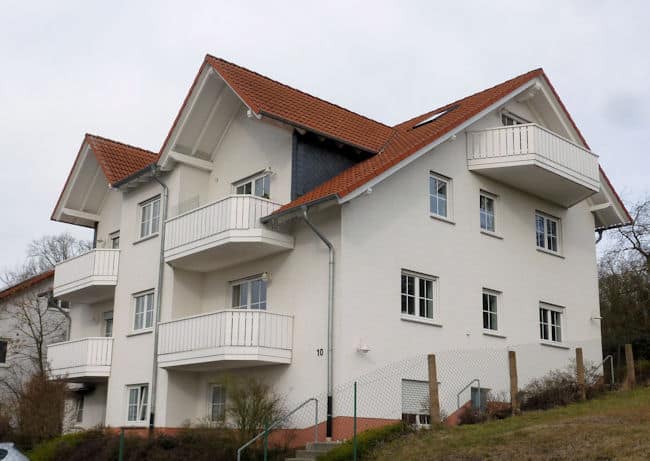 Das Bild zeigt ein dreistöckiges Wohnhaus mit Balkonen und rotem Ziegeldach an einem trüben Tag. Im Detail ist ein weißes, modernes Wohnhaus mit drei Stockwerken zu sehen. Das Haus hat ein rotes Ziegeldach und weiße Balkone mit Geländern. Die Fensterrahmen sind ebenfalls weiß. An der Fassade ist die Nummer "10" angebracht. Vor dem Haus befindet sich ein grüner Hang, der durch einen Maschendrahtzaun begrenzt wird. Eine Treppe führt zum Eingang des Hauses. Der Himmel ist grau und bewölkt, was auf einen trüben Tag hindeutet.