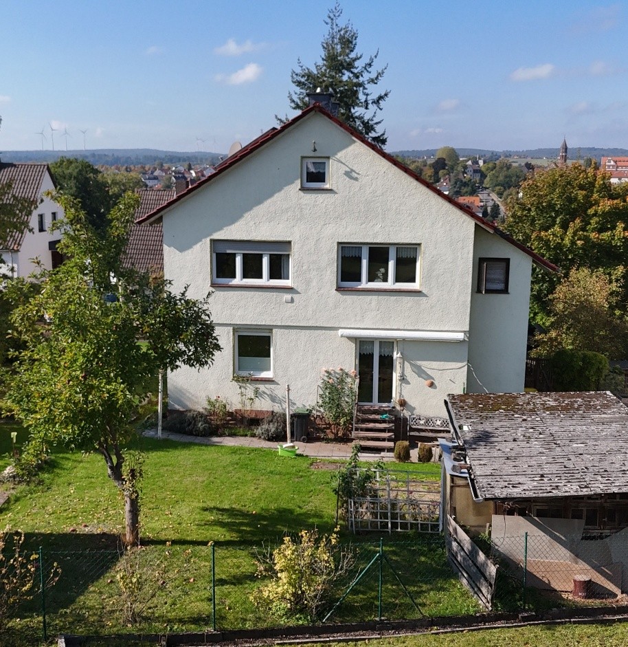 Das Bild zeigt ein zweistöckiges Einfamilienhaus mit einer hellbeigen Fassade und einem rotbraunen Ziegeldach. Das Haus befindet sich in einer ländlichen Umgebung mit viel Grün. Im Vordergrund ist ein gepflegter Rasen zu sehen, durch den ein Maschendrahtzaun verläuft. Auf dem Rasen stehen mehrere Bäume und Sträucher, einer davon, ein kleinerer Baum mit hellem Stamm, ist links im Bild platziert. Das Haus hat mehrere Fenster mit weißen Rahmen. Im Obergeschoss befinden sich links und rechts jeweils zwei nebeneinander liegende Fenster, im Erdgeschoss ein einzelnes, breiteres Fenster links und ein Eingangsbereich mit Glastür rechts, über dem eine weiße Markise ausgefahren ist. Über der Tür führt eine Treppe zu einer kleinen Terrasse. Rechts neben dem Haus ist ein kleinerer Anbau mit einem schrägen, mit grauen Dachziegeln gedeckten Dach zu erkennen, der an das Hauptgebäude anschließt. Im Hintergrund erstreckt sich eine hügelige Landschaft mit weiteren Häusern, Bäumen und in der Ferne sind einige Windräder sichtbar.