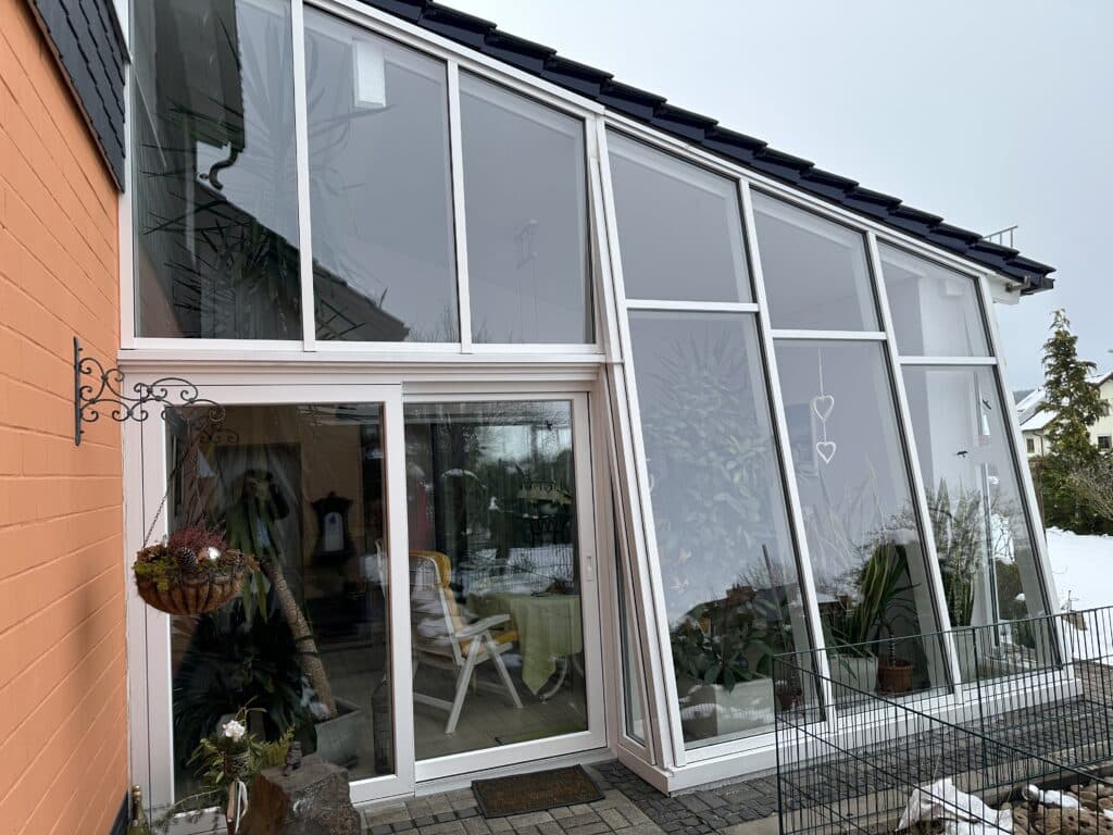 Das Bild zeigt eine modernen Wintergarten, der in ein orangefarbenes Haus angebaut ist. Der Wintergarten ist mit einem dunklen Ziegeldach bedeckt, das sich über die gesamte Länge erstreckt. Die Fenster sind groß und reichen vom Boden bis zum Dach, was viel natürliches Licht hereinlässt. Im Inneren des Wintergarten sind mehrere Pflanzen in verschiedenen Töpfen und Behältern zu sehen, darunter eine große Palme. Am Wintergarten befindet sich eine Tür die den Zugang zum Inneren ermöglicht. Direkt vor einem Glaselement hängt ein dekorativer Korb mit Pflanzen und dekorativen Gegenständen. Der Korb ist an einer kunstvoll geschmiedeten Metallhalterung an der Hauswand befestig.