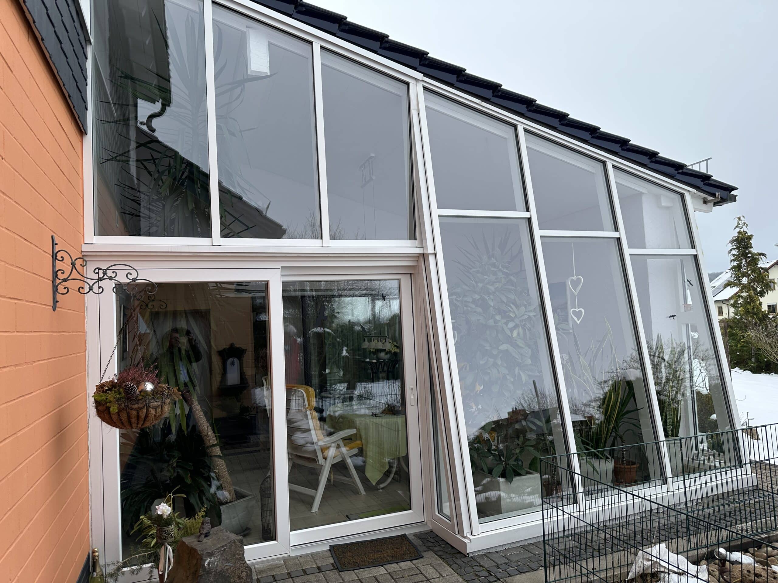 Das Bild zeigt eine modernen Wintergarten, der in ein orangefarbenes Haus angebaut ist. Der Wintergarten ist mit einem dunklen Ziegeldach bedeckt, das sich über die gesamte Länge erstreckt. Die Fenster sind groß und reichen vom Boden bis zum Dach, was viel natürliches Licht hereinlässt. Im Inneren des Wintergarten sind mehrere Pflanzen in verschiedenen Töpfen und Behältern zu sehen, darunter eine große Palme. Am Wintergarten befindet sich eine Tür die den Zugang zum Inneren ermöglicht. Direkt vor einem Glaselement hängt ein dekorativer Korb mit Pflanzen und dekorativen Gegenständen. Der Korb ist an einer kunstvoll geschmiedeten Metallhalterung an der Hauswand befestig.