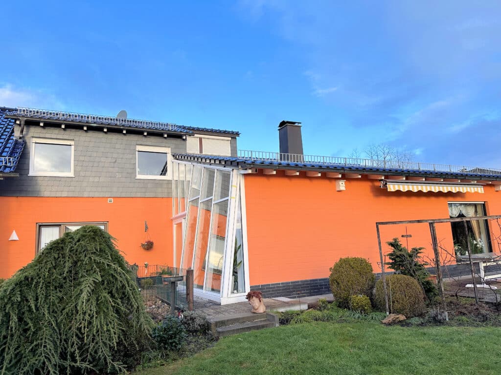 Die Aufnahme zeigt die Rückseite eines Einfamilienhauses mit einem umliegenden Garten. Das Haus hat eine auffällige orangefarbene Ziegelwand. Über der orangefarbenen Wand befindet sich ein graues Schieferdach, das mit einer weißen Bordüre verziert ist. Rechts ist ein Teil eines weiteren Gebäudes mit einem blauen Dach zu sehen. An der orangefarbenen Wand ist ein ausgefahrenes gelb-weiß gestreiftes Sonnensegel angebracht. Große Fenster spiegeln die umliegende Landschaft wieder. Der Garten ist größtenteils von einem grünen Rasen bedeckt. Im Vordergrund links befindet sich eine große, kugelförmige Zypresse, deren Äste herabhängen. Mehrere kleine Bäume und Büsche sind im Garten verteilt, einige davon sind kahler als andere. Der Himmel ist klar mit einigen weißen Wolken.