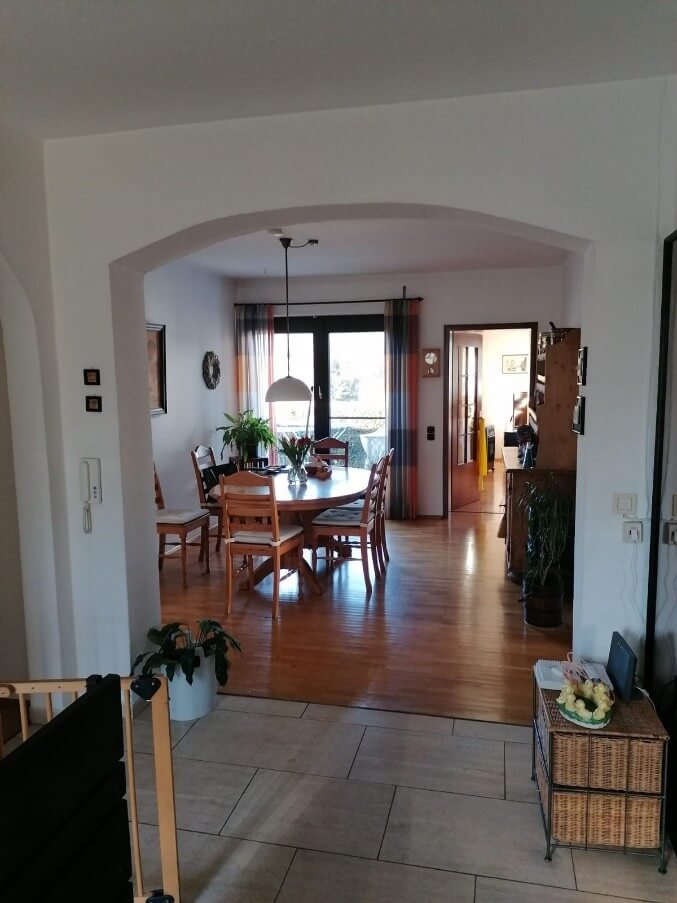 Die Aufnahme zeigt einen Durchgang zu einem Esszimmer und weiterführenden Räumen. Im Vordergrund befindet sich ein Teil eines Babygitters und eine Topfpflanze mit grünen Blättern in einem weißen Übertopf. Der Boden im Vordergrund ist mit hellbeigefarbenen Fliesen ausgelegt. Durch einen breiten, geschwungenen Durchgang gelangt man in das Esszimmer. Der Boden dort besteht aus glänzendem, hellem Parkett. In der Mitte des Raumes steht ein ovaler Esstisch aus Holz, an dem mehrere Holzstühle mit hellen Sitzkissen platziert sind. Auf dem Tisch steht eine Vase mit roten Blumen und eine Schale mit Obst oder Gemüse. Über dem Tisch hängt eine große, weiße Hängelampe. Hinter dem Esstisch befindet sich eine doppelflügelige Balkontür mit einem dunklen Rahmen, durch die Tageslicht einfällt und den Raum erhellt. An der Balkontür hängen farbige Vorhänge in Orange-, Rot- und Blautönen. An der Wand links des Durchgangs ist eine Freisprecheinrichtung für die Türanlage montiert. An der Wand im Esszimmer hängt links ein kreisförmiges Wandobjekt. Rechts im Hintergrund des Esszimmers ist eine offene Tür zu einem weiteren Raum zu sehen, durch die man in einen Gang mit gelbem Licht und Möbeln blickt. Ein weiteres Wandobjekt mit einem Zifferblatt, möglicherweise eine Uhr, hängt an der Wand im Esszimmer. Am rechten Bildrand, im Vordergrund des separaten Bereichs, steht ein niedriger Korpus aus geflochtenem Material mit zwei Schubladen auf einem Metallgestell. Darauf befindet sich ein ein Arrangement aus gelben Objekten, vielleicht Dekoration. Weiter hinten im rechten Bereich des Bildes steht ein hoher, dunkler Holzschrank, der teilweise mit Pflanzen dekoriert ist.