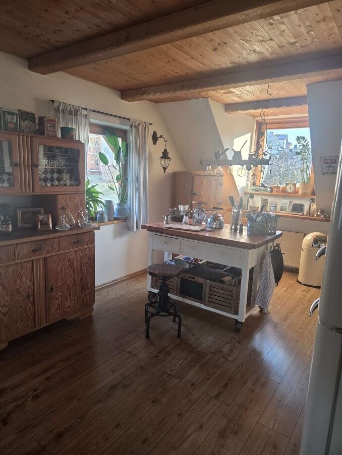 Die Aufnahme zeigt eine gemütliche, rustikale Küche mit viel Holz. Im Vordergrund dominiert ein großzügiger, hölzerner Küchenschrank mit Glastüren und Schubladen. Seine Oberfläche ist mit verschiedenen Gegenständen wie Flaschen, Krügen und Gläsern dekoriert. Die Türen des Schranks geben den Blick auf Regale frei, auf denen in karierter Weise angeordnete Gläser oder Behälter zu sehen sind. Daneben befindet sich ein Fenster mit gestreiften Vorhängen, hinter dem grüne Pflanzen stehen und das Tageslicht hereinlässt. Im mittleren Bereich des Bildes steht eine helle Kücheninsel mit einer Holzplatte und mehreren Schubladen. Auf der Insel sind diverse Küchenutensilien wie Töpfe, Waagen und Messer angeordnet. Davor steht ein kleiner, schwarzer Hocker mit einem runden Sitz. Unter der Kücheninsel befinden sich Holzkisten mit Gegenständen. Der Boden der Küche ist mit breiten, braunen Holzdielen ausgelegt, die ein warmes Ambiente schaffen. Die Decke ist ebenfalls mit Holz verkleidet und mit sichtbaren Holzbalken versehen, was den rustikalen Charakter des Raumes unterstreicht. Im Hintergrund sind weitere Elemente der Küche zu erkennen, darunter ein weiteres Holzmöbelstück, ein Fenster mit Blick nach draußen auf eine leicht unscharfe ländliche Szene, und am rechten Bildrand ein Teil eines hellen Kühlschranks und ein cremefarbener Mülleimer.