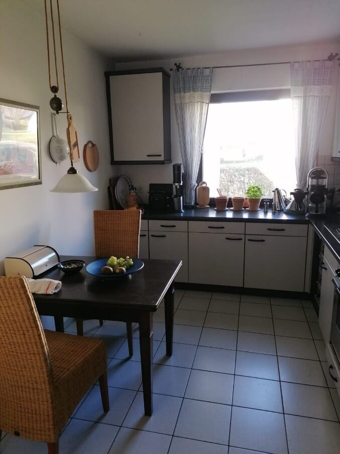 Die Aufnahme zeigt eine helle und gemütliche Küche im Tageslicht. Im Vordergrund steht ein dunkelbrauner Holztisch, der von zwei geflochtenen Stühlen umgeben ist. Auf dem Tisch befindet sich eine blaue Schale mit einer grünen Traube und einem kleinen Obststück sowie eine weiße Brotdose und ein kleiner schwarze Schüssel mit etwas darin. Die Stühle haben eine Sitzfläche aus hellem Geflecht. Der Boden ist mit hellgrauen quadratischen Fliesen ausgelegt. Die Wand im Hintergrund ist weiß gestrichen. An der Wand hängen mehrere Küchenutensilien, darunter ein Sieb, ein Holzbrett und ein weiteres Holzgegenstand. Eine Pendelleuchte mit einem weißen Schirm hängt von der Decke herab. Links neben dem Tisch hängt ein gerahmtes Bild an der Wand, dessen Inhalt jedoch nicht klar erkennbar ist. Im mittleren Bereich der Küche befindet sich eine Küchenzeile mit weißen Unterschränken und einer dunklen Arbeitsplatte. Über den Unterschränken sind Geräte wie eine Kaffeemaschine, ein Wasserkocher und ein weiteres Küchengerät aufgestellt. An der Wand über der Arbeitsplatte ist ein grauer Hängeschrank angebracht. Ein großes Fenster befindet sich hinter der Küchenzeile und ist mit einem gestreiften Vorhang in Blau- und Weißtönen sowie mit Spitzen verziert. Durch das Fenster fällt helles Tageslicht, das einen Teil des Raumes beleuchtet und Schatten wirft. Auf der Fensterbank stehen mehrere kleine Blumentöpfe mit grünen Pflanzen und zwei Holzfiguren.