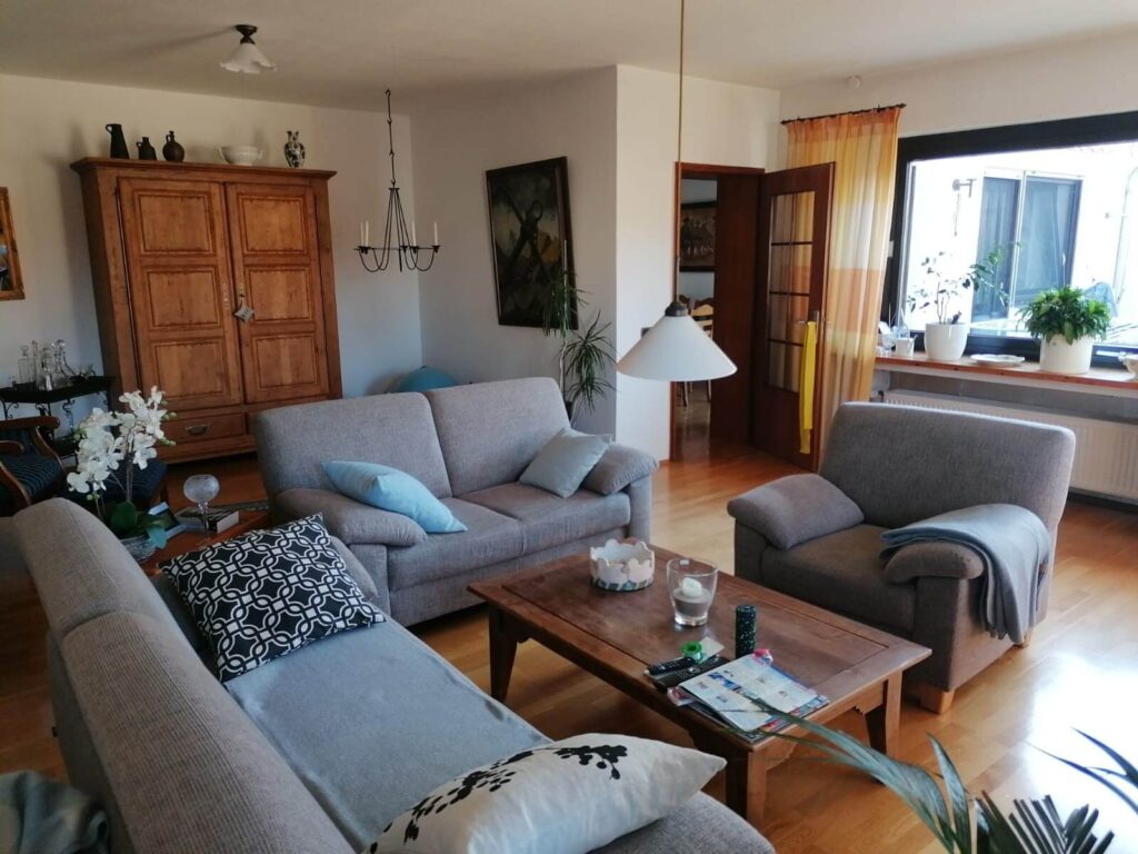 Das Bild zeigt ein gemütliches Wohnzimmer mit einem rustikalen Charme. Im Vordergrund befindet sich eine hellgraue Couchgarnitur, bestehend aus einer langen, L-förmigen Couch und einem separaten Sessel. Die Couch ist mit mehreren Kissen dekoriert, darunter zwei hellblaue Kissen, ein Kissen mit einem geometrischen Muster in Schwarz und Weiß und ein weiteres Kissen mit einem schwarzen Muster auf weißem Grund. Auf dem Sessel liegt eine hellblaue Decke. Der Couchtisch ist aus dunklem Holz gefertigt und mit verschiedenen Gegenständen belegt: eine kleine Schale mit einer weißen Dekoration, mehrere Fernbedienungen und Zeitschriften. An der Seite des Tisches sind einige Pflanzen zu sehen, deren Blätter in Richtung des Betrachters wachsen. Im Hintergrund links steht ein großer, hölzerner Schrank im Landhausstil mit zwei Türen und einer Schublade darunter. Auf dem Schrank sind mehrere dunkle Vasen und eine weiße Schale platziert. Daneben hängt eine dunkle, mehrarmige Kerzenleuchter-Lampe von der Decke. An der Wand über dem Schrank hängt ein rechteckiges Gemälde mit einem dunklen Rahmen, das eine düstere Szene darstellt. Links neben dem Gemälde und über der Couch, an der weißen Wand, ist eine weitere Deckenlampe mit einem weißen Lampenschirm zu sehen. Rechts im Bild befinden sich ein großes Fenster mit Blick nach draußen und eine Tür mit Glaseinsätzen, die in einen anderen Raum führt. Durch die Tür ist ein Esstisch mit Stühlen erkennbar. Vor dem Fenster stehen zwei Blumentöpfe mit grünen Pflanzen auf einer Fensterbank, über der eine Heizung angebracht ist. Der Fensterbereich wird von gelb-orange gestreiften Vorhängen umrahmt.