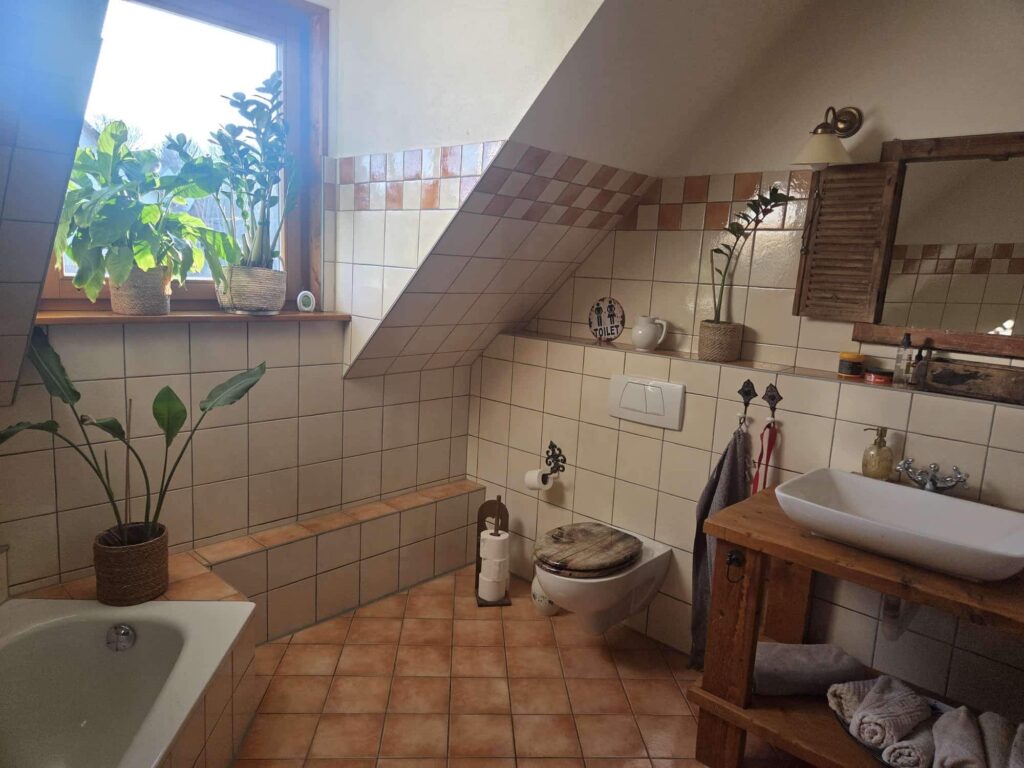 Das Bild zeigt ein Badezimmer mit Dachschräge. Der Boden ist mit großen, terracottafarbenen Quadratfliesen ausgelegt. Die Wände sind im unteren Bereich ebenfalls mit diesen Fliesen verkleidet, während der obere Bereich und die Dachschräge mit cremefarbenen Quadratfliesen versehen sind, die in regelmäßigen Abständen von Streifen aus hellbraunen und cremefarbenen Fliesen durchzogen sind. Im linken Teil des Bildes befindet sich eine weiße Badewanne, die teilweise von links aus dem Bild herausragt. Auf dem Rand der Badewanne, der ebenfalls gefliest ist, stehen zwei Topfpflanzen. Die größere Pflanze links hat breite, grüne Blätter und steht in einem geflochtenen Korb. Die kleinere Pflanze rechts hat schlanke, aufrechte Blätter und steht ebenfalls in einem geflochtenen Korb. Vor dem Fenster steht noch eine dritte Pflanze mit großen, elegant geschwungenen grünen Blättern in einem braunen Korb auf dem gefliesten Badewannenrand. Ein großes Fenster mit Holzeinfassung befindet sich im hinteren linken Bereich des Raumes und lässt viel helles Tageslicht herein. Durch das Fenster sind schemenhaft Bäume und der Himmel zu erkennen. An der linken Seite des Fensters hängt ein grauer Vorhang. Weiter rechts im Bild steht eine weiße Toilette mit einem rustikalen Holzdeckel. Direkt neben der Toilette steht auf dem Boden ein dunkler Metallständer mit drei Rollen Toilettenpapier. Über der Toilette an der Wand hängt ein kleines Schild mit der Aufschrift "TOILET" und zwei stilisierten Personenfiguren. Daneben steht eine weiße Krug. Der Hauptbereich des Raumes wird von einem Waschbecken dominiert, das auf einem rustikalen Holztisch steht. Das Waschbecken ist weiß, langgezogen und hat eine glänzende Oberfläche. Die Armatur ist aus Metall mit zwei Drehgriffen. An der Wand hinter dem Waschbecken befinden sich hängende Handtücher. Links vom Waschbecken hängt ein dunkelgraues Handtuch an einem Haken und daneben ein rotes Band an einem weiteren Haken. Rechts vom Waschbecken ist ein Spender für Flüssigseife in einem bernsteinfarbenen Behälter zu sehen. Der Holztisch unter dem Waschbecken hat zwei Ablageflächen. Auf der unteren Ablage liegen ordentlich zusammengefaltete Handtücher, hauptsächlich in Grau- und Beigetönen. Hinter dem Waschbecken und der Seifenschale sind einige kleinere Behälter und Flaschen zu sehen, die wahrscheinlich Kosmetikartikel enthalten. Über dem Waschbecken hängt ein großer Spiegel mit einem breiten, dunklen Holzrahmen. Links neben dem Spiegel ist eine braune Holzjalousie angebracht, die anscheinend zu einem Schrank gehört oder als Dekoration dient. Über der Jalousie und dem Spiegel ist eine Wandleuchte mit einem cremefarbenen Lampenschirm und einem messingfarbenen Gestell befestigt.