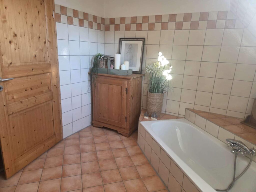 Das Bild zeigt ein Badezimmer, das rustikal und einladend wirkt. Im Vordergrund befindet sich eine freistehende Badewanne, die mit cremefarbenen Fliesen umrahmt ist. Der Wasserhahn ist glänzend metallisch und hat einen flexiblen Schlauch. Daneben stehen zwei kleine Segelboote als Dekoration. Links neben der Wanne steht ein gewebter Korb mit einer Topfpflanze, deren helle Blüten das Sonnenlicht reflektieren und fast weiß leuchten. Hinter der Pflanze befindet sich ein niedriges, abgestuftes Podest, das ebenfalls mit den cremefarbenen Fliesen verkleidet ist und die Wanne umgibt. Im mittleren Bereich des Bildes steht ein altes, holzfarbenes Möbelstück. Auf diesem Möbelstück befindet sich ein geflochtener Behälter, in dem eine grüne Pflanze mit langen, herabhängenden Blättern arrangiert ist. Daneben steht eine kleine, hölzerne Holzkiste, die mit einem Griff versehen ist und in der drei weiße Kerzen unterschiedlicher Höhe stehen. Hinter den Kerzen hängt ein gerahmtes Bild im Vintage-Stil, das eine junge Frau in einem Kleid darstellt. Ganz links im Bild ist eine Holztür mit sichtbarer Holzmaserung zu sehen. Die Tür ist leicht geöffnet und hat einen silbernen Türgriff. Der Boden im gesamten Badezimmer ist mit quadratischen, terracottafarbenen Fliesen ausgelegt. Die Wände sind überwiegend mit weißen, quadratischen Fliesen gefliest. Oberhalb der weißen Fliesen verläuft ein dekoratives Band mit abwechselnden braunen und weißen Kacheln, das sich bis zur Decke fortsetzt. Die Decke ist schräg und hell.