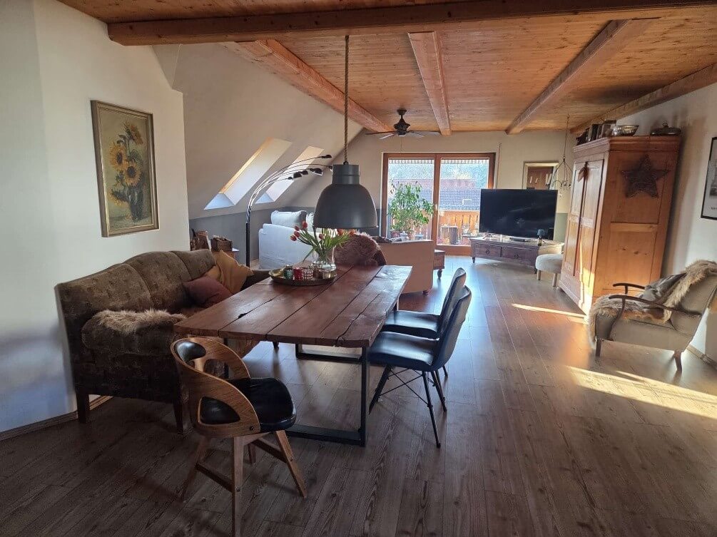 Das Bild zeigt ein gemütliches Ess- und Wohnzimmer im Dachgeschoss. Der Raum ist von hellem Tageslicht durchflutet, das durch Dachfenster und eine große Glastür fällt und lange Schatten auf den Holzboden wirft. Die Decke ist mit Holzbalken verziert, die dem Raum eine rustikale Note verleihen. Im Vordergrund dominiert ein massiver Holztisch mit einer dunklen Metallkonstruktion als Tischbein. Auf dem Tisch steht eine Vase mit roten Tulpen und ein Tablett mit Kerzen. An den Tisch grenzen vier Stühle: zwei schwarze Stühle mit Metallbeinen, die eher modern wirken, und ein rustikalerer Holzstuhl mit schwarzen Sitzflächen, der links im Bild zu sehen ist. Links im Bild steht ein braunes, gemustertes Sofa, auf dem sich einige Kissen und eine flauschige Decke befinden. An der Wand über dem Sofa hängt ein gerahmtes Gemälde mit Sonnenblumen. Der weitere Raum ist in verschiedene Bereiche unterteilt. Im Hintergrund ist eine offene Wohnlandschaft angedeutet, mit einem hellen Sofa und einer Stehlampe mit mehreren Armen. Eine Glastür führt auf einen Balkon oder eine Terrasse mit Blick auf Bäume und Häuser. Ein Fernsehgerät steht auf einem dunklen Holzmöbelstück. Rechts im Bild steht ein hoher, alter Holzschrank, der mit verschiedenen Objekten dekoriert ist, darunter eine Sternform. Daneben befindet sich ein Sessel, der mit einer flauschigen Decke bedeckt ist und ebenfalls von den Sonnenstrahlen beleuchtet wird.