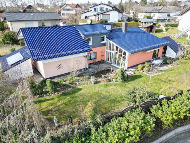 Das Bild zeigt eine Luftaufnahme eines Wohnhauses mit Garten. Das Haus ist in mehrere Abschnitte unterteilt. Ein Teil hat ein schlichtes, hellbraunes Äußeres mit rechteckigen Öffnungen, die wie Fenster oder Lüftungsschlitze aussehen. Daneben befindet sich ein größerer Teil des Hauses mit einer leuchtend korallenroten Fassade, der in ein großes, schräg geneigtes Glas-Erkerfenster übergeht. Dieses Glasdach ist mit einem blauen Ziegeldach bedeckt, das sich über den größten Teil des Hauses erstreckt. Ein kleinerer Teil des Hauses, der an den korallenroten Abschnitt angrenzt, ist graubraun und hat ebenfalls ein blaues Ziegeldach mit einem kleinen Schornstein. An einem der Dächer sind auch mehrere Solarpaneele installiert, die hellblau-silbern glänzen. Der Garten ist üppig und grün, mit Rasenflächen, Büschen und Bäumen. Im Vordergrund befindet sich ein dichter Bewuchs mit dunkelgrünen Blättern und einigen kahlen Ästen. Mehrere kleine, geschwungene Bäume oder Sträucher stehen auf dem Rasen. Hinter dem korallenroten Haus erstreckt sich eine gepflasterte Terrasse mit weißen Gartenmöbeln und einem Wäscheständer. Ein kleiner Teich ist im Garten links vom Glas-Erkerfenster zu sehen. Am Rand des Grundstücks verläuft ein niedriger, dunkler Holzzaun, der von üppigem Grün überwachsen ist. Im Hintergrund sind weitere Wohnhäuser mit verschiedenen Dachformen und Fassaden zu erkennen, die sich auf einem sanften Hügel verteilen. Der Himmel ist klar und hellblau, was auf einen sonnigen Tag hindeutet.