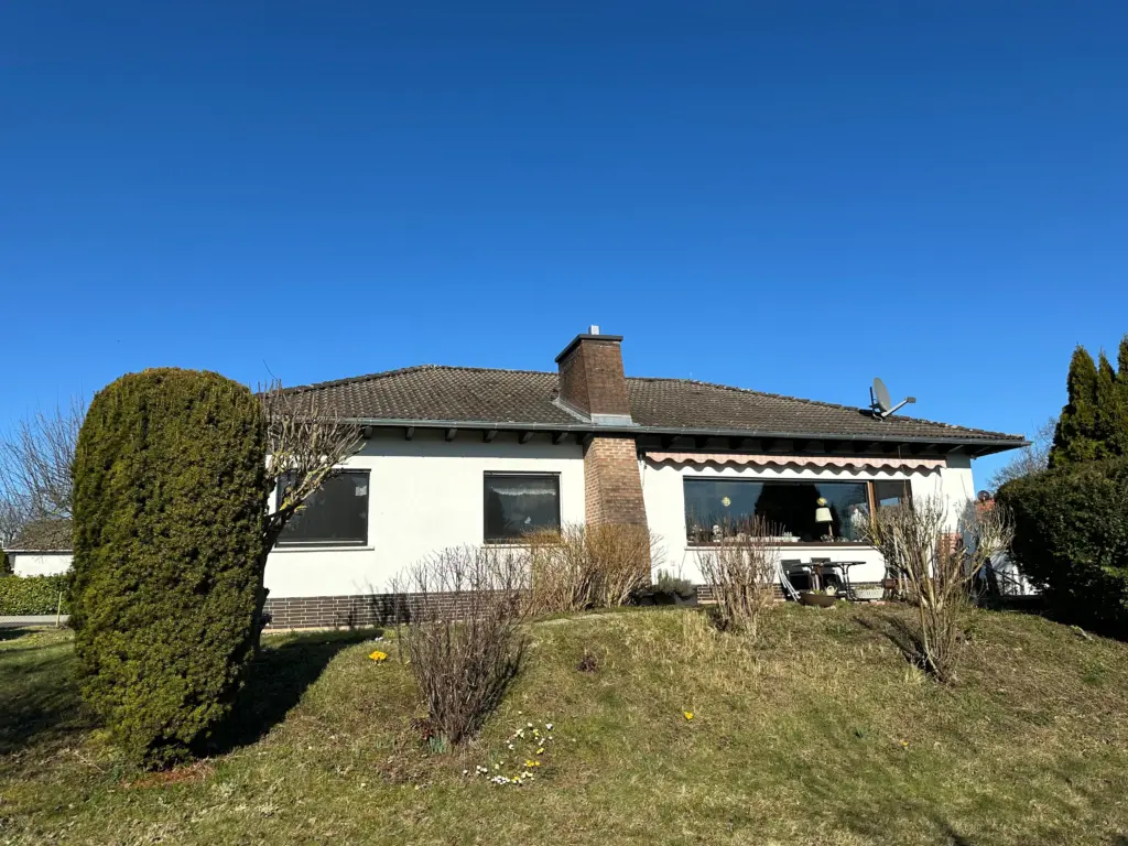 Das Bild zeigt ein freistehendes Einfamilienhaus an einem sonnigen Tag. Der Himmel ist strahlend blau und wolkenlos. Das Haus ist einstöckig, mit einem dunkelgrauen Ziegeldach und weißen Wänden. An der Vorderseite befinden sich mehrere Fenster; eines davon ist ein großes Panoramafenster, durch das man einen Blick ins Innere werfen kann, wo ein Tisch mit Stühlen, eine Lampe und weitere Gegenstände zu erkennen sind. Neben dem Haus steht ein markanter, dichter und hochgewachsener grüner Koniferenbusch. Vor dem Haus erstreckt sich ein leicht abfallender, von Gras bewachsener Hang mit einigen spärlichen Büschen und kleineren, kahlen Sträuchern. Vereinzelt blühen kleine gelbe und weiße Frühlingsblumen, wie zum Beispiel Narzissen. Ein gemauerter Schornstein ragt aus dem Dach hervor und auf der rechten Dachseite ist eine Satellitenschüssel montiert. Die Schatten im Vordergrund deuten auf eine helle Sonneneinstrahlung hin. Die Komposition des Bildes ist so gewählt, dass das Haus und sein direkter Vorgartenbereich im Vordergrund stehen, während der Himmel den oberen Teil dominiert. Im Hintergrund links sind weitere Häuser und Bäume zu erkennen. Die Farbpalette ist geprägt von kräftigem Blau des Himmels, dem Grün der Konifere, dem Grau des Daches, dem Weiß der Hauswände und den erdigen Tönen des Hanges und der Vegetation.
