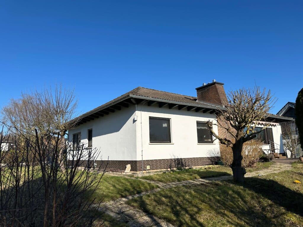 Das Bild zeigt ein einstöckiges weißes Haus mit einem braunen Ziegelsockel. Das Haus hat ein dunkles, geneigtes Dach mit sichtbaren Dachsparren. An der rechten Seite des Hauses befindet sich ein Ziegelkamin. Vor dem Haus erstreckt sich ein gepflegter Garten mit grünen Rasenflächen und schattenspendenden Bäumen. Einige der Bäume sind noch kahl, da es sich um eine frühe Jahreszeit handeln könnte. Die Fenster des Hauses sind dunkel getönt und spiegeln den klaren blauen Himmel wider. Im Vordergrund des Bildes sind niedrig wachsende Büsche und verschlungene Äste von Bäumen zu sehen, die Schatten auf den Rasen werfen. Der Boden ist teilweise mit Kieswegen durchzogen. Das Licht ist hell und die Schatten sind scharf, was auf einen sonnigen Tag hindeutet. Die Farben sind satt und kontrastreich, dominiert von dem strahlenden Blau des Himmels und den verschiedenen Grüntönen des Grases. Die Komposition ist leicht nach rechts geneigt, wobei das Haus den Hauptfokus bildet und der Garten sich drumherum entfaltet.