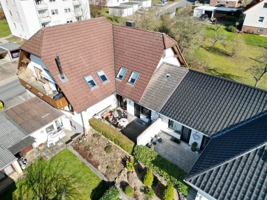Das Bild zeigt eine Luftaufnahme eines Wohnhauses und seines Gartens. Das Haus ist weiß gestrichen und hat mehrere Dächer mit unterschiedlichen Dachziegeln. Ein Teil des Daches ist rot und besteht aus Ziegeln, während ein anderer Teil grau und mit Schieferplatten gedeckt ist. An den Dachflächen befinden sich mehrere Dachfenster. Vor dem Haus befindet sich ein kleiner Vorgarten mit einem gepflegten Rasen und einer Steinmauer. Darauf folgen mehrere Beete mit unterschiedlichen Pflanzen, darunter Sträucher und kleine Bäume. Weiter hinten im Garten ist ein größerer Rasenbereich zu sehen, der von Bäumen gesäumt wird. Am rechten Rand des Bildes sind weitere Häuser und Gebäude zu erkennen. Das Bild ist von oben nach unten ausgerichtet, wobei das Haus den größten Teil des Bildes einnimmt. Die Farben sind hauptsächlich natürliche Töne: rot und grau für die Dächer, weiß für die Hauswände, grün für den Rasen und die Pflanzen und braun für die Erde und die Bäume. Im Bereich der Terrasse vor dem Haus sind einige Gartenmöbel zu sehen, darunter ein Tisch und Stühle. Ein kleinerer Anbau mit einem Pultdach aus Wellblech befindet sich links am Haus. Rechts vom Hauptgebäude erstreckt sich ein weiterer Gebäudeteil mit dunklen Dachziegeln. Die Aufnahme vermittelt einen Eindruck von einem gepflegten Wohnhaus in einer ruhigen Nachbarschaft.
