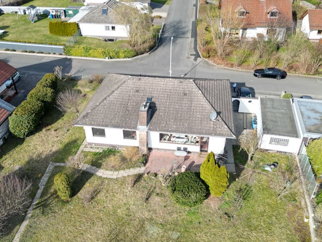 Das Bild zeigt eine Luftaufnahme eines Wohngebiets an einem sonnigen Tag. Im Vordergrund und im Zentrum des Bildes befindet sich ein weißes Einfamilienhaus mit einem grauen Ziegeldach. Das Haus hat mehrere Fenster und einen Schornstein. Vor dem Haus erstreckt sich eine Terrasse aus rötlichen Ziegelsteinen, die von einer niedrigen Mauer umgeben ist. Der Garten vor dem Haus ist größtenteils mit Gras bewachsen, durchzogen von Wegen aus Steinplatten. In der rechten unteren Ecke des Bildes ist ein kleiner Baum mit leuchtend gelben Blättern zu sehen, daneben ein dunkelgrüner, kugelförmiger Busch. Weiter hinten im Garten stehen vereinzelte Bäume und Sträucher, die noch kahl sind, was auf eine frühlingshafte Jahreszeit hindeutet. Im mittleren Bereich des Bildes, rechts vom Haupthaus, befindet sich eine Garage mit flachem, grauem Dach. Daneben sind weitere kleinere Gebäude mit ähnlichen Dächern zu erkennen. Eine schwarze Limousine parkt vor einer Einfahrt, die zu einem der Gebäude führt. Im Hintergrund erstreckt sich eine asphaltierte Straße, die sich leicht bergauf schlängelt. Entlang der Straße sind weitere Häuser zu sehen, einige mit roten Ziegeldächern und weißen Fassaden, andere mit grauen Dächern. Die Häuser sind von Grünflächen und Bäumen umgeben. Am oberen linken Bildrand sind weitere Häuser mit Gärten und einem Spielgerüst zu erkennen.