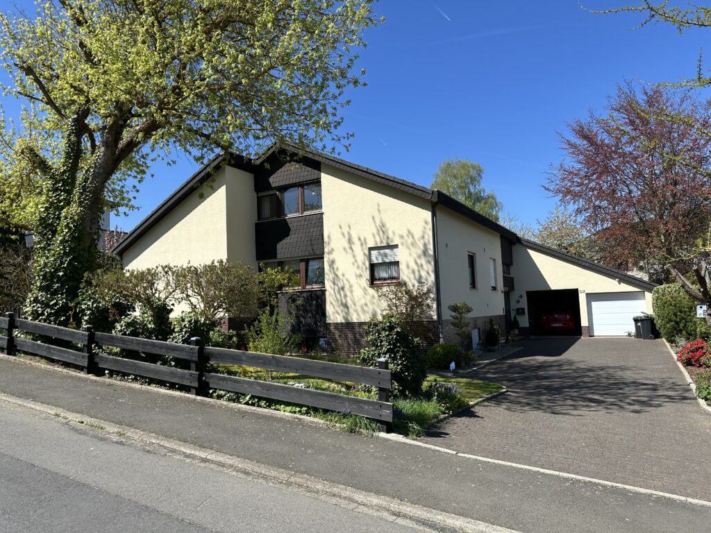 Die Aufnahme zeigt ein großzügiges Einfamilienhaus an einem sonnigen Tag. Das Haus ist an der Fassade in einem hellen Beigeton gehalten und hat ein dunkles, schräg abfallendes Dach. Ein Teil der Fassade, der sich im oberen Bereich befindet, ist dunkel verkleidet und von mehreren rechteckigen Fenstern unterbrochen. Vor dem Haus erstreckt sich ein Garten, der von einem niedrigen, dunklen Holzzaun abgegrenzt wird. Im Vordergrund verläuft ein asphaltierter Weg, der zu einem gepflasterten Vorplatz führt. Der Zaun ist aus dunklem Holz gefertigt und mit mehreren senkrechten Pfosten verstärkt. Davor und dahinter ist üppige grüne Vegetation zu sehen, darunter auch Efeu, das an den Zaunpfosten und die Hauswand klettert. Der Garten ist mit verschiedenen Sträuchern und kleinen Bäumen bepflanzt, einige davon blühen zart. Im Bereich des Zauns sind auch kleine lila Blüten zu erkennen. Links im Bild ragt ein großer Baum mit hellem, frisch ausgetriebenem Laub auf, dessen Äste weit in den Himmel reichen. Sein Stamm ist dicht mit Efeu bewachsen. Im Hintergrund des Hauses sind weitere Bäume zu sehen, einer mit rötlichem Laub und ein weiterer mit hellem, lockerem Blätterwerk. Rechts vom Haus befindet sich eine Garagenzufahrt mit einem elektrischen Garagentor, das in einem hellen Grau gehalten ist. In der geöffneten Einfahrt ist ein rotes Auto zu erkennen. Neben dem Garagentor steht eine dunkle Mülltonne.