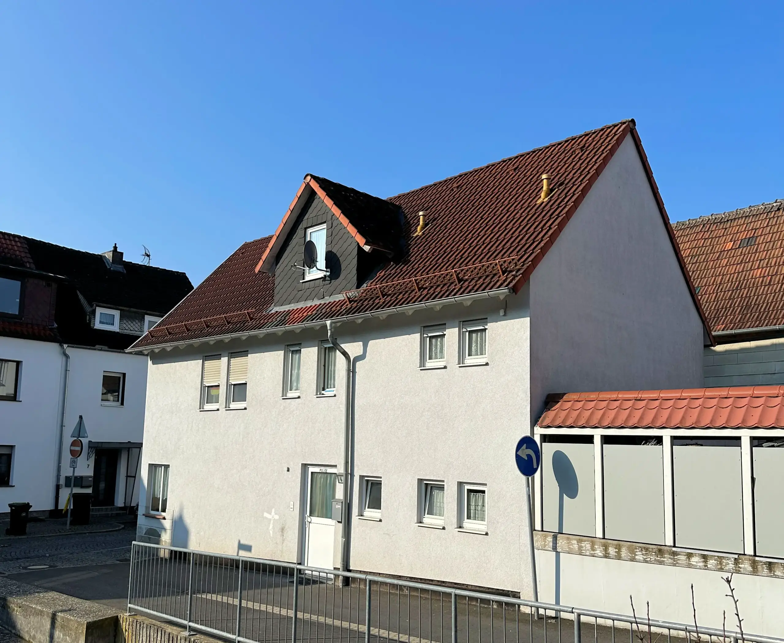 Auf dem Bild ist ein zweigeschossiges gepflegtes Wohngebäude mit nach rechts angebauten eingeschossigen Anbau zu sehen. Das Gebäude hat eine helle Fassade. Das Dach ist mit roten Ziegelsteinen eingedeckt. In der Mitte des Daches befindet sich eine Gaube mit Fenster und SAT-Spiegel. Die Fenster sind weiß ebenso die Hauseingangstür. Im Obergeschoss befinden sich sechs Fenster im Erdgeschoss vier. Vor dem Wohnhaus befindet sich eine gepflasterte Straßenfläche welche zum Nachbargrundstück mit einem Zaun gesichert ist. Im Hintergrund und links der Immobilie sind weitere Gebäude erkennbar. Der Himmel ist blau und wolkenlos.