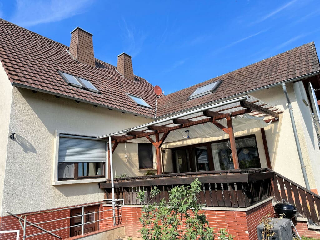 Auf dem Bild sieht man die Terrasse eines Wohnhauses. Die Terrasse ist überdacht. Das Geländer ist aus dunklem Holz. Die Farbe der Fassade ist in einen hellen Gelbton. Das Dach ist mit einer Ziegeleindeckung versehen. Auf der linken Dachhälfte erkennt man zwei Schornsteine, eine Satellitenschüssel und drei Dachfenster. Ein weiteres Dachfenster befindet sich auf der rechten Dachhälfte. Der untere Bereich des des Gebäudes ist mit roten Klinkersteinen versehen. Auf der linken Seite erkennt man eine Treppengeländer und ein Tür, was auf einen Kellerzugang hindeutet. Über der Kellertür befindet sich ein Fenster, dass Rollo ist halb nach unten gelassen. Auf der rechten Seite befindet sich der Treppenaufgang zur Terrasse vor der Treppe steht ein schwarzer Kugelgrill und eine graue Aufbewahrungsbox. Der Himmel ist blau mit ein paar leichten Schleierwolken.
