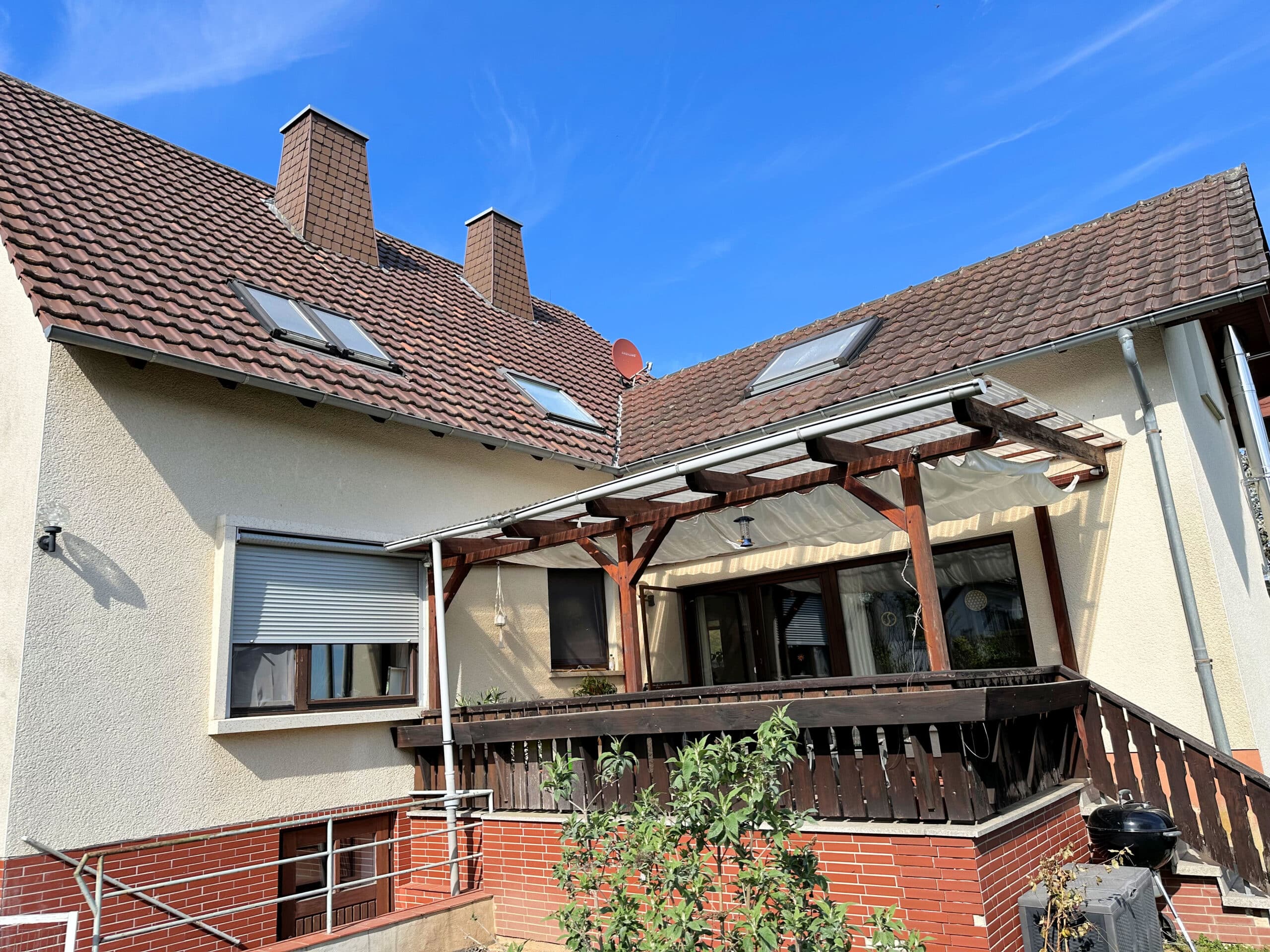 Auf dem Bild sieht man die Terrasse eines Wohnhauses. Die Terrasse ist überdacht. Das Geländer ist aus dunklem Holz. Die Farbe der Fassade ist in einen hellen Gelbton. Das Dach ist mit einer Ziegeleindeckung versehen. Auf der linken Dachhälfte erkennt man zwei Schornsteine, eine Satellitenschüssel und drei Dachfenster. Ein weiteres Dachfenster befindet sich auf der rechten Dachhälfte. Der untere Bereich des des Gebäudes ist mit roten Klinkersteinen versehen. Auf der linken Seite erkennt man eine Treppengeländer und ein Tür, was auf einen Kellerzugang hindeutet. Über der Kellertür befindet sich ein Fenster, dass Rollo ist halb nach unten gelassen. Auf der rechten Seite befindet sich der Treppenaufgang zur Terrasse vor der Treppe steht ein schwarzer Kugelgrill und eine graue Aufbewahrungsbox. Der Himmel ist blau mit ein paar leichten Schleierwolken.
