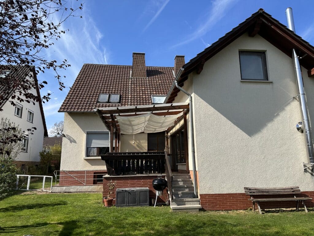 Das Bild zeigt eine Rückansicht eines Hauses an einem sonnigen Tag. Im Vordergrund erstreckt sich ein grüner Rasen, der von der tiefstehenden Sonne beleuchtet wird, was zu einigen Schatten führt. Links ist ein Ast eines Baumes mit wenigen dunkelroten Blättern zu sehen, der sich von einem strahlend blauen Himmel mit vereinzelten weißen Schleierwolken abhebt. Das Haus ist in zwei Teile gegliedert. Der linke Teil hat eine weiße Fassade und ein dunkelbraunes Ziegeldach. Auf dem Dach sind zwei kleine Dachfenster zu erkennen. Darunter befinden sich zwei rechteckige Fenster mit weißen Fensterläden. Daneben ist ein weiteres Gebäude mit einer helleren Fassade zu sehen, das aber größtenteils im Schatten liegt. Der rechte Teil des Hauses hat eine hellbeige, grob verputzte Fassade und ebenfalls ein dunkelbraunes Ziegeldach. An der Dachkante sind dunkle Holzelemente angebracht. Ein rostfarbener Sockel aus Ziegeln verläuft entlang der unteren Wand. An der rechten Außenwand des Hauses ist ein langer, glänzender Edelstahlschornstein montiert, der fast bis zur Dachkante reicht. Darunter ist ein einzelnes rechteckiges Fenster mit dunklem Rahmen zu sehen, dessen Glas die Umgebung spiegelt. Vor dem rechten Teil des Hauses befindet sich eine Terrasse, die von einer Holzkonstruktion mit einem weißen, wellenförmig drapierten Sonnensegel überspannt wird. Von der Terrasse führen graue Steinstufen mit einer Kante aus rotem Ziegelstein in den Garten hinunter. Links neben den Stufen steht ein schwarzer Kugelgrill mit einem Deckel und einem Untergestell, daneben ein grauer Kunststoffschrank. Auf dem Sockel des Hauses, links neben dem Grill, steht ein kleiner brauner Blumentopf mit einem vertrockneten Pflänzchen. Am Ende der Stufen und leicht nach rechts versetzt steht eine einfache Holzbank, die aus zwei nebeneinanderliegenden Sitzflächen besteht und auf mehreren Metallbeinen ruht. Die Bank steht auf dem grünen Rasen und wirft einen deutlichen Schatten auf die Wand des Hauses.