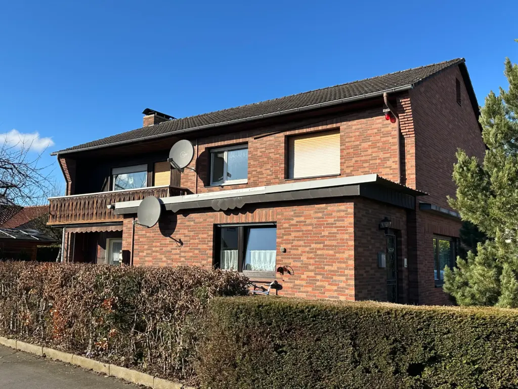 Das Bild zeigt ein zweistöckiges Ziegelhaus mit einem dunklen Ziegeldach. Vor dem Haus erstreckt sich ein dichter, brauner Heckenstreifen, der von einem grauen Betonrand und einem schmalen Streifen Asphalt oder Weg begrenzt wird. Der Himmel ist strahlend blau und wolkenlos. Die Fassade des Hauses ist aus rötlich-braunen Ziegelsteinen gebaut, die in einem typischen Mauerverband verlegt sind. Im Erdgeschoss sind rechts ein Fenster mit weißen Gardinen und links ein weiteres Fenster, ebenfalls mit Gardinen, zu sehen. Über diesem Fenster befindet sich ein dunkles Vordach mit geschwungenem Rand. Das Obergeschoss weist eine markante Balkonkonstruktion auf, die mit einem geschnitzten Holzvorsprung verziert ist. An der Fassade sind zwei graue Satellitenschüsseln angebracht, eine über dem Balkon und eine darunter. Links am Haus befindet sich ein Schornstein, der aus der Dachlinie ragt. An der rechten Ecke des Hauses, im Obergeschoss, sind zwei rote Blitzleuchten an der Regenrinne montiert. Daneben ragt ein dichter Baum mit nadeligen Zweigen ins Bild.
