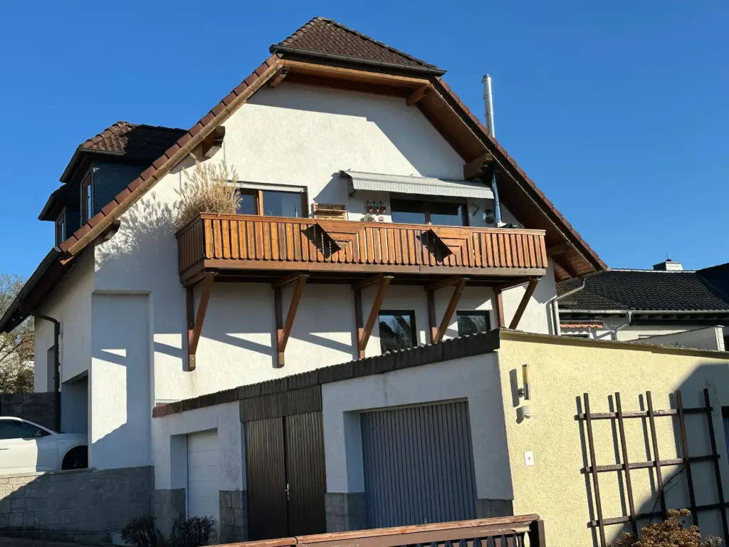 Das Bild zeigt ein modernes Einfamilienhaus mit einem Balkon und einer Garage an einem sonnigen Tag. Der Himmel ist strahlend blau und bildet einen starken Kontrast zur weißen Fassade des Hauses. Das Dach ist mit braunen Ziegeln gedeckt und hat eine ausgeprägte Neigung. Das auffälligste Merkmal des Hauses ist der hölzerne Balkon im ersten Stock. Er ist mit vertikalen Holzlatten und diagonalen Stützen versehen, die ihm ein rustikales Aussehen verleihen. Auf dem Balkon stehen ein paar Pflanzen, darunter hohe, trockene Gräser, die im Wind wedeln. Über dem Balkon befindet sich eine ausfahrbare Markise in hellen Streifen. An der Fassade des Hauses sind mehrere Fenster zu sehen, die dunkle Rahmen haben und die Reflexionen des blauen Himmels und der umliegenden Bäume zeigen. Ein Teil des Hauses ist mit einer grauen, steinartigen Verkleidung versehen, insbesondere im Bereich der Garage. Unter dem Balkon befindet sich ein Garagenanbau mit zwei Toren. Das linke Tor ist dunkelbraun und hat eine vertikale Lamellenstruktur. Das rechte Tor ist grau und besteht aus horizontalen Sektionen. Die Garage ist teilweise mit einer hellgelben Wand verkleidet, an der eine Wandleuchte und ein Rankgitter angebracht sind. Im Vordergrund des Bildes ist ein Teil eines braunen Holzzauns zu erkennen. Links neben dem Haus parkt ein weißes Auto, dessen hinterer Teil sichtbar ist.