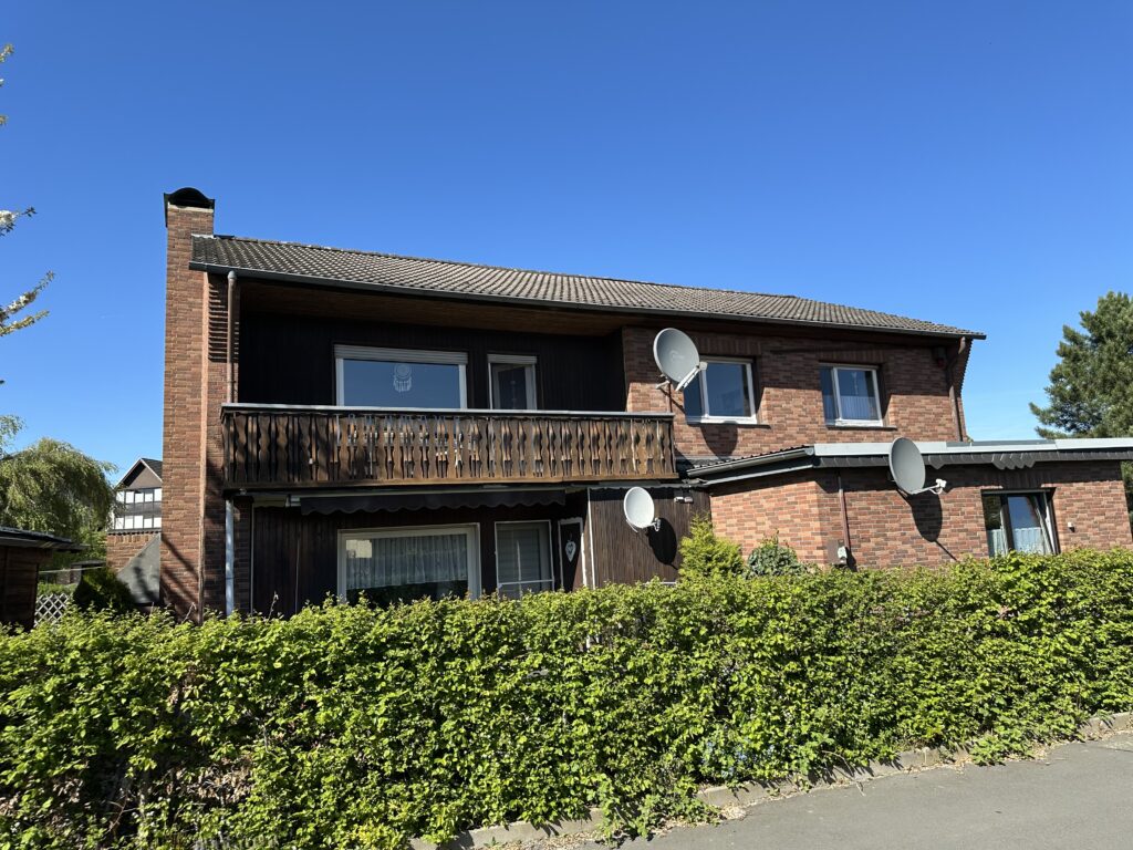 Auf dem Bild ist ein verklinkertes Wohnhaus mit zwei Vollgeschossen, Satteldach und Balkon zu sehen. Links am Gebäude ist ein Schornstein angebaut. Im rechten Bereich des Hauses befindet sich ein eingeschossiger Anbau mit Fenster und Flachdach. Des Weiteren sind an der Fassade drei SAT-Anlagen angebracht. Die Fenster im Haupthaus sind weiß. Im Erdgeschoss befindet neben dem Anbau eine Terrasse. Vor der Immobilie zur Straße hin ist eine Buchenhecke sichtbar. Der Himmel ist strahlend blau.