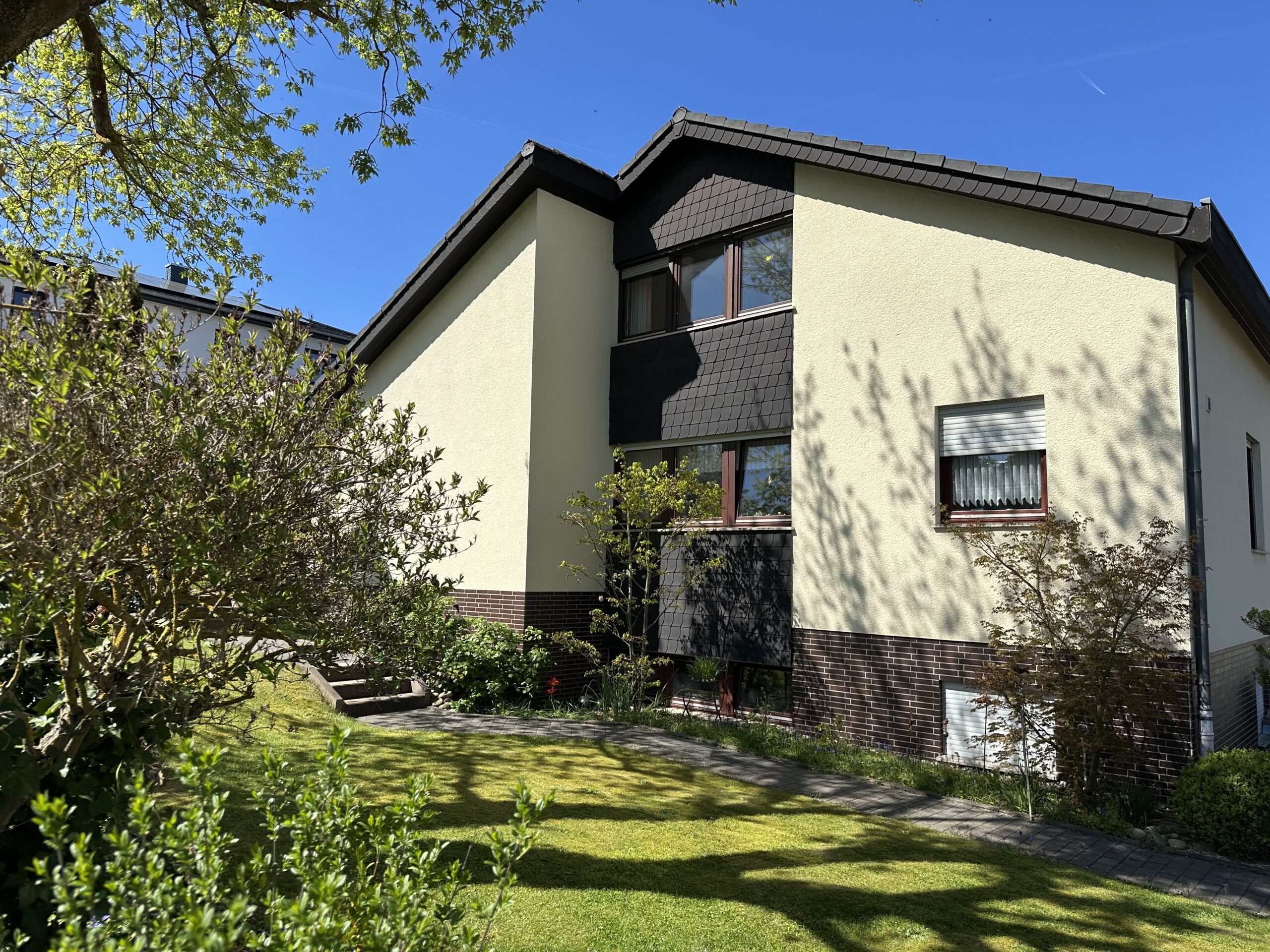 Das Bild zeigt ein zweistöckiges Haus an einem sonnigen Tag. Die Fassade des Hauses ist hellgelb verputzt und mit dunkelbraunen Schindeln an den Giebeln und an der Dachlinie verziert. Der untere Teil des Hauses ist mit dunkelroten Ziegeln verkleidet. Es gibt mehrere Fenster mit braunen Rahmen. Einige Fenster sind mit weißen Vorhängen zu sehen. Auf der linken Seite des Hauses ragt ein großer Baum mit hellgrünen Blättern ins Bild. Im Vordergrund befindet sich ein gepflegter Rasen, der von Sonnenlicht durchflutet ist und Schatten von Bäumen wirft. Ein gepflasterter Weg führt durch den Garten. Rechts neben dem Haus wachsen Büsche und kleinere Bäume.