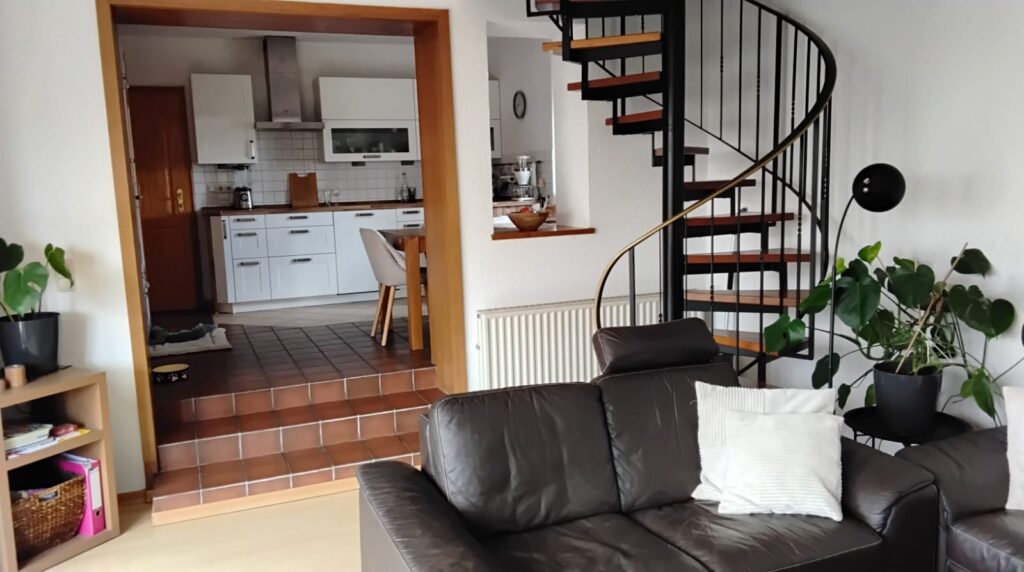 Diese Bild zeigt einen Blick aus dem Wohnzimmer in eine Küche. Im Vordergrund des Bildes sieht man einen dunkelbraunen Ledersessel. Hinter dem Ledersessel erstreckt sich eine Wendeltreppe die nach oben führt. Neben der Treppe sind eine Stehlampe und eine grüne Pflanze in einem anthrazitfarbenen Blumentopf. In der Mitte des Bildes sieht man einen Durchgang in die Küche. Die Küche erreicht man über zwei geflieste Stufen. Die Fliesen sind in einem Braunton und erstrecken sich über den kompletten Küchenfußboden. Die Küche auf die man blickt hat weise Fronten und eine dunkle Arbeitsplatte. Eine Dunstabzugshaube befindet sich zwischen zwei Hängeschränken.