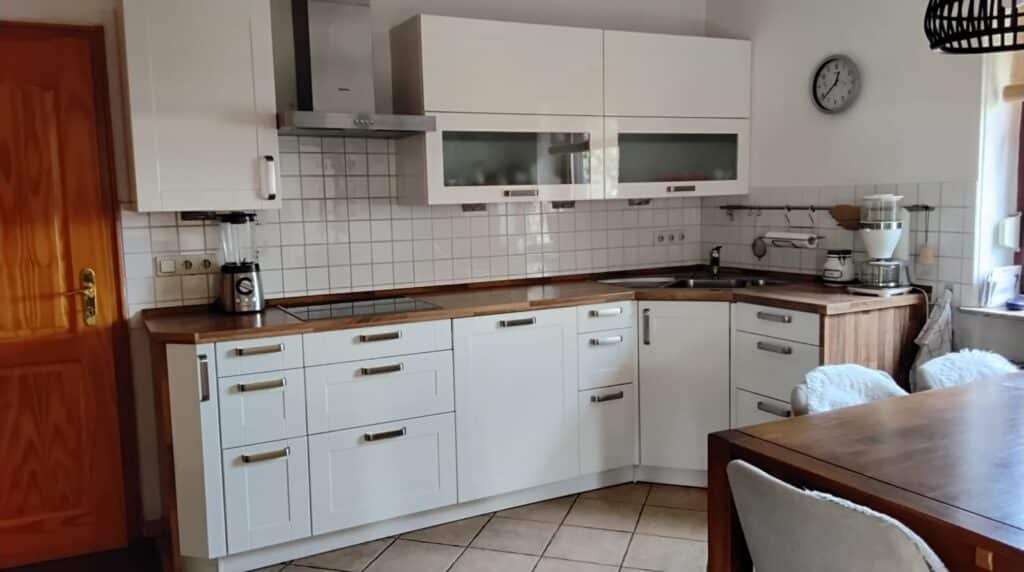 Das Bild zeigt eine helle und moderne Küche. Der Hauptfokus liegt auf den weißen Küchenschränken, die in einer L-Form angeordnet sind. Die Oberschränke haben teilweise Glastüren, durch die man einige Gegenstände erkennen kann. Die Arbeitsplatte besteht aus hellem Holz, was einen schönen Kontrast zu den weißen Fronten bildet. Linkerhand im Bild ist eine hölzerne Tür mit goldenen Beschlägen zu sehen. Darauf folgend im Arbeitsbereich steht ein silberner Standmixer auf der Arbeitsplatte. Die Wand hinter der Arbeitsplatte ist mit weißen, rechteckigen Fliesen verkleidet, die ein Schachbrettmuster ergeben. Eine Edelstahlabzugshaube ist über dem Kochfeld angebracht. Rechts im Bild schließt sich die Küchenzeile an. Dort befinden sich ein Spülbecken, Armatur und ein weißer Kaffeemaschine auf der Arbeitsplatte. Eine Wanduhr mit silbernem Rahmen und schwarzen Ziffern hängt an der weißen Wand über dem Spülbereich. Daneben sind einige Küchenutensilien an einer Stange aufgehängt, darunter ein Papierrollenhalter und ein Geschirrtuch. Im Vordergrund ist ein Teil eines großen Holztisches zu sehen, der mit zwei Stühlen belegt ist. Die Stühle sind mit hellen, flauschigen Bezügen versehen. Der Tisch und die Stühle sind in einem warmen Holzton gehalten. Die Beleuchtung im Raum ist hell und natürlich, was durch ein Fenster rechts im Bild angedeutet wird. Die Komposition ist so gewählt, dass sie einen guten Überblick über die Küchenzeile und den Essbereich gibt. Die Aufnahme ist leicht von oben und schräg gemacht.