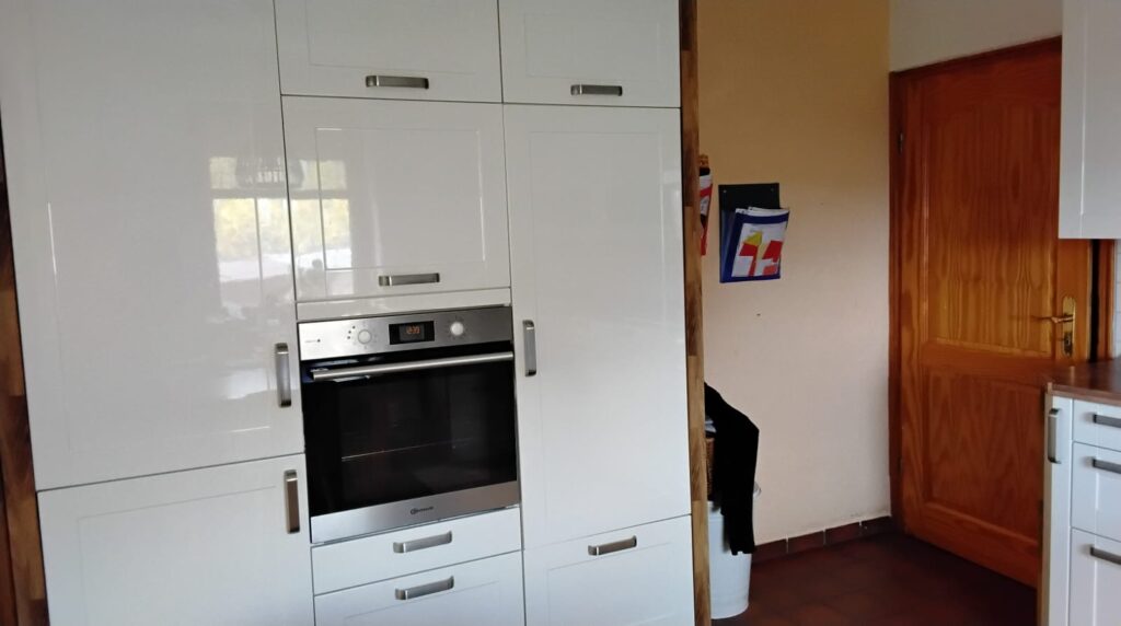 Das Bild zeigt eine Küche mit modernen weißen Hochglanzschränken, die an der Wand angebracht sind. In den Schrank integriert ist ein Einbauofen aus Edelstahl mit einem digitalen Display, das die Uhrzeit 12:39 anzeigt. Über dem Ofen befinden sich weitere Schranktüren mit silbernen Griffen. Links neben dem Ofen sind die Schränke hochglänzend weiß, und man kann Reflexionen von Fenstern und möglicherweise der Außenwelt erkennen, was auf eine helle Umgebung hindeutet. Rechts von den weißen Küchenschränken befindet sich eine Holztür aus hellem Holz mit einer deutlichen Maserung und einem goldenen Türgriff. Die Tür ist geschlossen und steht im Kontrast zu den modernen weißen Küchenmöbeln. An der Wand neben der Tür hängt ein blauer Zeitschriftenständer mit verschiedenen farbigen Blättern oder Magazinen. Die Wand selbst ist in einem warmen, hellen Orangeton gestrichen. Vor der Wand und neben den Küchenschränken steht ein weißer Mülleimer, teilweise verdeckt von einem schwarzen Kleidungsstück, das über einen Korb gehängt ist. Der Boden ist mit dunkelroten oder braunen Fliesen ausgelegt.