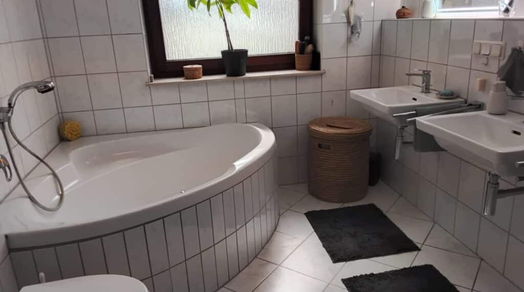 Das Bild zeigt ein Badezimmer. Im Vordergrund links ist eine weiße, eckige Badewanne mit geschwungenen Rändern zu sehen. Sie ist teilweise mit kleinen, hellgrauen Fliesen verkleidet. An der Wand über der Wanne befinden sich größere, quadratische, weiße Fliesen mit grauen Fugen. Ein Duschkopf mit Schlauch ist seitlich an der Wanne angebracht. Links neben der Wanne liegt ein gelber Badeschwamm. Im Hintergrund des Bildes befindet sich ein Waschbeckenbereich mit zwei nebeneinander angeordneten, weißen Waschbecken. Beide Waschbecken sind schlicht und modern gestaltet und haben verchromte Armaturen. Unter den Waschbecken sind silberne Rohre und Handtuchhalter zu erkennen. Zwischen den beiden Waschbecken und links davon steht ein runder Wäschekorb aus hellem, geflochtenem Material mit einem Deckel. Daneben liegen zwei dunkelgraue Teppiche auf dem hell gefliesten Boden. An der Wand über den Waschbecken sind weitere weiße Fliesen angebracht. Rechts oben im Bild ist ein Fenster mit einer Milchglasscheibe und einem dunkelbraunen Rahmen zu sehen. Auf der Fensterbank steht eine Topfpflanze mit grünen Blättern in einem schwarzen Topf, ein kleiner runder Behälter aus geflochtenem Material und ein weiterer Behälter, der Haarbürsten enthält. Auf der Fensterbank rechts befinden sich weitere Dekorationsgegenstände. Die Beleuchtung im Raum ist hell und gleichmäßig, was auf Tageslicht durch das Fenster und/oder künstliche Beleuchtung schließen lässt. Die Farbpalette des Badezimmers ist überwiegend hell, mit viel Weiß und Grau, akzentuiert durch das Braun des Fensterrahmens und des Wäschekorbs sowie das Gelb des Schwamms und das Grün der Pflanze.