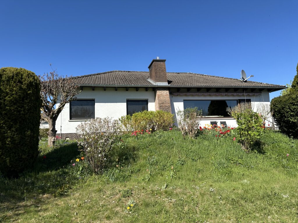 Das Bild zeigt ein freistehendes Einfamilienhaus an einem sonnigen Tag. Der Himmel ist strahlend blau und wolkenlos. Das Haus ist einstöckig, mit einem dunkelgrauen Walmdach und weißen Fassaden. An der Südseite befinden sich mehrere Fenster; eines davon ist ein großes Panoramafenster mit Balkontür, sowie zwei weitere Fenster. Vor dem Panoramafenster befindet sich eine Terrasse mit Bestuhlung. Neben dem Haus steht ein markanter, dichter und hochgewachsener grüner Koniferenbusch. Vor dem Objekt erstreckt sich ein leicht abfallender, von Gras bewachsener Hang mit gepflegten Büschen und kleineren Sträuchern. Vereinzelt blühen hier Frühlingsblumen, wie zum Beispiel Narzissen und Tulpen. Ein gemauerter und verklinkerter Schornstein ragt aus dem Dach hervor und grenzt an die Terrasse in westlicher Richtung. Auf der rechten Dachseite ist eine Satellitenanlage montiert.