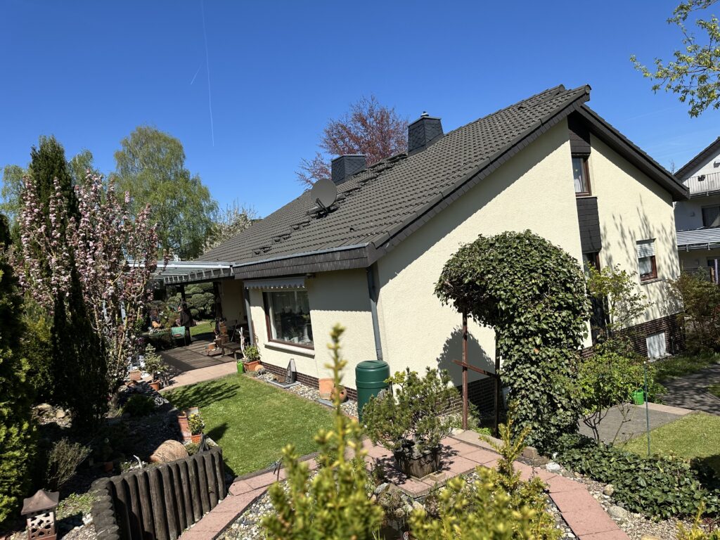 Das Bild zeigt ein sonniges Tageslicht und zeigt die Außenansicht eines Hauses und seines Gartens. Im Vordergrund sind verschiedene Pflanzen und kleine Bäume mit grünen Nadeln zu sehen, die unscharf sind und den Fokus auf das Haus und den Garten lenken. Darunter befindet sich ein niedriger, hölzerner Zaun. Der Garten ist gut gepflegt und verfügt über einen sattgrünen Rasen, der von Kieswegen mit roten Steineinfassungen begrenzt wird. Auf den Kieswegen sind verschiedene Topfpflanzen und dekorative Elemente wie ein großer, terrakottafarbener Topf und eine stilisierte Laterne zu sehen. Links im Bild, am Rande des Rasens, steht eine grüne Regentonne. Ein Teil des Hauses ist sichtbar, mit einer hellgelben Fassade und einem dunklen, geneigten Ziegeldach. An der Fassade sind braune Fensterrahmen und ein dunkler Kamin zu erkennen. Auf dem Dach sind eine Satellitenschüssel und zwei dunkle Schornsteine montiert. An der Fassade befinden sich auch einige dunkelbraune Zierelemente. Rechts von der Haustür gibt es eine durch eine Markise abgedeckte Terrasse mit Gartenmöbeln, die von einer üppig bewachsenen Säule aus Efeu eingerahmt wird. Im Hintergrund sind weitere Bäume und Sträucher zu sehen, darunter ein blühender Baum mit rosa Blüten links im Bild und ein Baum mit rotem Laub weiter hinten. Der Himmel ist strahlend blau.