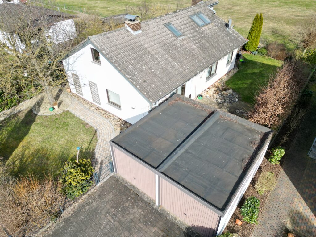 Die Aufnahme zeigt eine Drohnenaufnahme eines Wohnhauses mit einem angrenzenden Garagenbau. Das Hauptgebäude ist weiß verputzt und hat ein graues Ziegeldach, auf dem zwei Dachfenster und ein Schornstein zu sehen sind. Vor dem Haus erstreckt sich ein gepflegter Rasen, der von einem geschwungenen Weg aus hellbraunen Pflastersteinen durchzogen wird. Am linken Bildrand steht ein kahler Baum mit verschlungenen Ästen, dessen Schatten auf den Rasen fällt. Daneben steht ein grüner Ball auf einem Topf. Das Garagengebäude hat eine braune Fassade und ein flaches, dunkles Dach, das mit einer Bitumenbahn gedeckt ist und deutlich verschmutzt aussieht. Davor befindet sich eine Auffahrt, ebenfalls mit grauen Pflastersteinen belegt. Entlang der Grenzen des Grundstücks sind Büsche und Hecken zu erkennen, die größtenteils noch keine Blätter tragen hindeutet. Im Hintergrund ist eine weite Grünfläche zu sehen, die von einigen Bäumen und einem Zaun begrenzt wird.