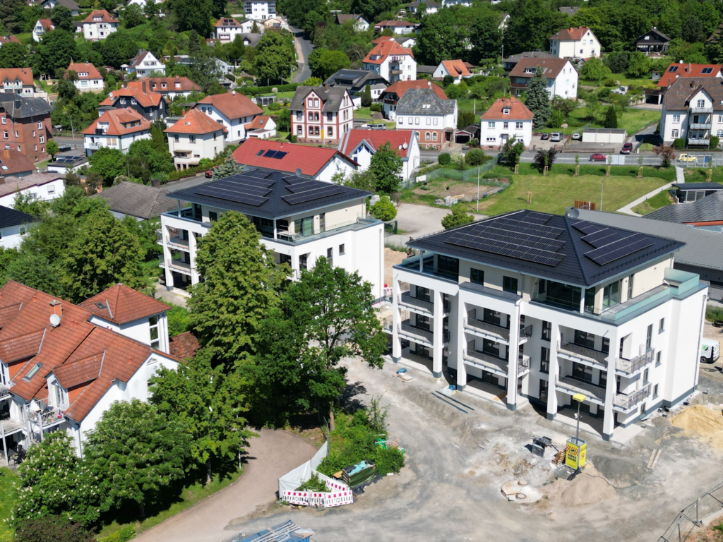 Auf dieser Drohnenaufnahme sind zwei moderne nach Süden ausgerichtete Mehrfamilienhäuser mit dunklen Dachziegel und Photovoltaikanlagen zu sehen. Jede der jeweils 11 Wohnungen pro Haus verfügt über sehr große Balkone und bodentiefe Fenster. Im Obergeschoss befinden sich sehr große überdachte Dachterrassen mit Glasgeländern. Die Außenanlage der Wohnanlage ist noch nicht fertig gestellt. Hier befinden sich noch Bauzäune und Werkzeug. Vor dem linken Gebäude ist eine Videoüberwachung in Form eines Turmes zu sehen. In direkter Nachbarschaft befindet sich ebenfalls ein Mehrfamilienhaus mit roten Dachziegel. Im Hintergrund der modernen Gebäude erkennt man weitere Häuser mit Gärten, Grünanlagen, sowie eine Straße mit Fahrzeugen.