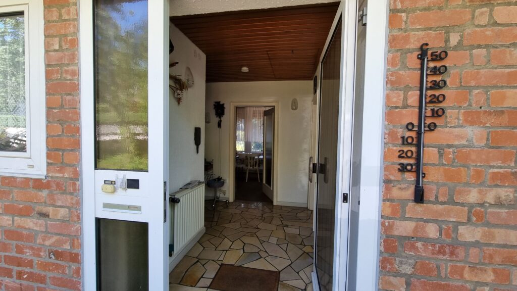 Das Bild zeigt den Eingangsbereich eines Hauses mit einer offenen Tür, die den Blick in einen Flur freigibt. Vor dem Haus, an einer Ziegelwand befestigt, hängt ein schwarzes Thermometer mit deutlichen Markierungen, das etwa 27 Grad Celsius anzeigt. Die Ziegelwand ist in warmen Rottönen gehalten. Links von der Eingangstür befindet sich ein Fenster mit weißem Rahmen. Die Eingangstür selbst ist ebenfalls weiß umrahmt und hat ein großes Milchglasfenster, durch das man verschwommen grüne Vegetation erkennt. An der Tür sind eine Klingel, ein Schalter und ein Briefschlitz angebracht. Der Flur ist mit unregelmäßig geformten Steinplatten in verschiedenen erdigen Tönen ausgelegt. An der weißen Wand links hängt eine weiße Heizkörper. Daneben sind verschiedene Gegenstände dekoriert, darunter eine Kuckucksuhr und eine Art Wanddekoration mit verschiedenen Elementen. Weiter im Flur ist eine weitere Tür zu sehen, die zu einem Zimmer führt, in dem ein Tisch und Stühle erkennbar sind und durch ein Fenster Tageslicht einfällt. Die Decke des Flurs ist mit Holzpaneelen verkleidet.