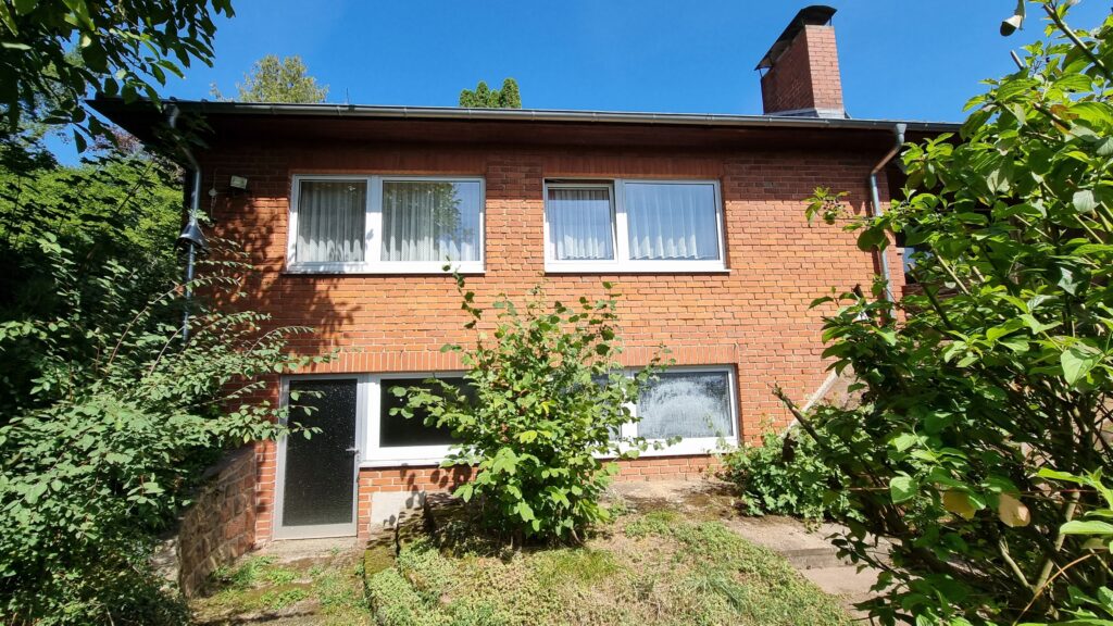 Das Bild zeigt die Fassade eines roten Backsteinhauses an einem sonnigen Tag. Der Himmel ist klar und strahlend blau. Die Fassade ist in einem warmen Rotton gehalten, die einzelnen Ziegel sind gut erkennbar. Das Haus hat mehrere weiße Fensterrahmen, einige davon mit zarten, weißen Vorhängen. Im oberen Bereich des Hauses befinden sich zwei größere Fenster nebeneinander, darunter ein kleineres Fenster und daneben eine dunkle Tür mit einer Milchglasscheibe. Vor dem Haus wachsen üppige grüne Büsche und kleinere Bäume. Die Vegetation ist dicht und bildet einen starken Kontrast zur roten Ziegelwand. Einzelne Blätter und Zweige reichen in das Bild hinein und verdecken teilweise die Hauswand und die Fenster. Der Boden vor dem Haus ist leicht uneben und mit Gras und kleineren Pflanzen bewachsen. Man erkennt einige graue Steine, die wohl Teil einer kleinen Mauer oder eines Weges sind. Am oberen rechten Rand des Hauses ragt ein gemauerter Schornstein aus roten Ziegeln in den blauen Himmel. An der Hauswand sind an einigen Stellen Regenrinnen und ein Scheinwerfer zu sehen.