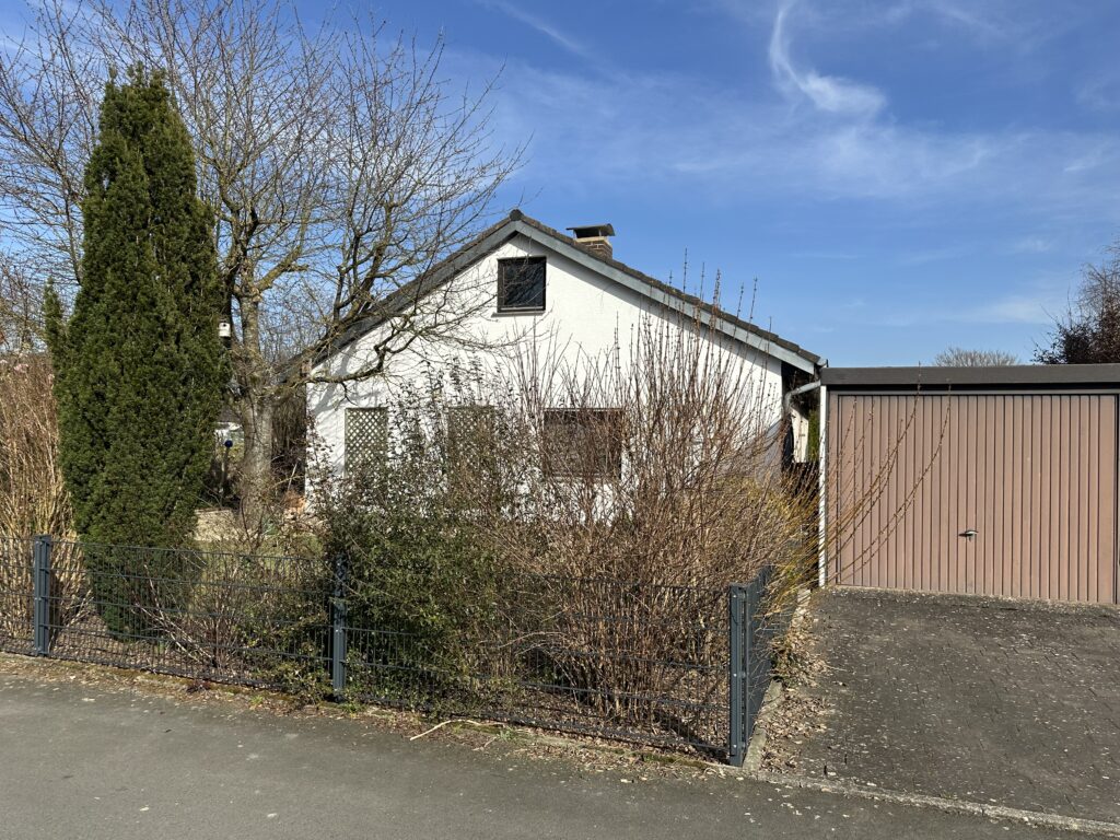 Auf dem Bild ist ein etwas zurückgesetztes eingeschossiges Wohnhaus mit Satteldach und Schornstein zu sehen. Rechts neben dem Haus befindet sich eine Garage mit Garagentor. Die Fläche vor der Garage ist bis zur asphaltierten Straße gepflastert. Das Grundstück vor dem Haus ist eingezäunt und mit Sträuchern und einem circa 4 Meter hohen, schlanken Nadelbaum bewachsen. Im linken Bereich vor dem Hauses ein großer Baum sichtbar. Dieser hat noch keine Blätter ebenso die Sträucher vor dem Haus. Man kann den Giebel des Hauses gut erkennen. im Dachgeschoss befindet sich ein Fenster, im Erdgeschoss ebenfalls und zusätzlich erkennt man zwei Rankgitter. Der Himmel ist strahlend blau.