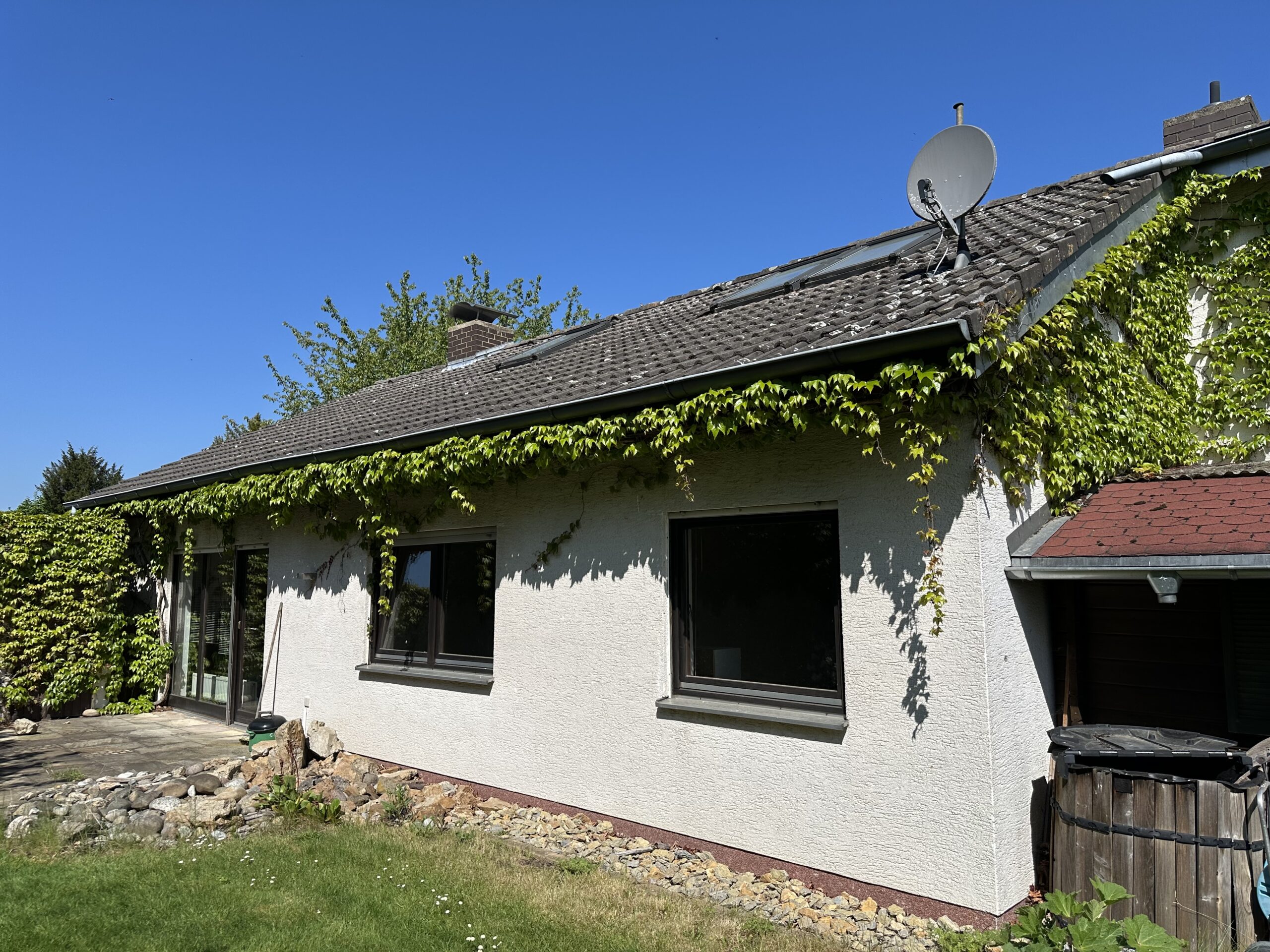 Auf diesem Foto ist die Südseite und ein kleiner Teil der Giebelseite eines Bungalows abgebildet. Auf dieser Längsseite sind zwei Fenster und ein bodentiefes Terrassenfenster mit Terrassentür zu sehen. Die Fassade ist weiß und der Sockelanstrich rotbraun. Die Dacheindeckung ist dunkel. Im Dach sind drei Dachflächenfenster eingebaut und ein SAT-Spiegel installiert. Eberfalls sind zwei Schornsteine zu erkennen. Auf dem rechten Bildrand ist direkt an der Giebelseite ein kleines mit roten Schindeln eingedecktes Vordach zu sehen. Unter diesem Dach befindet sich eine Wassertonne. Über ein Teil der Giebelseite und entlang der Längsseite im Bereich Dachrinne wächst Wilder-Wein. Entlang der Hauswand befindet sich ein Kiesbett und mündet im hinteren Bereich in eine Terrasse. Der Himmel ist blau, ohne Wolken.