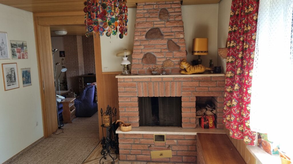 Das Bild zeigt ein gemütliches Wohnzimmer mit einem auffälligen Kamin im Mittelpunkt. Der Kamin ist aus rotem Backstein gebaut und mit größeren, unregelmäßigen Steinen verziert. Über dem Kamin, an der Wand, hängen zwei Lampen. Die linke Lampe hat einen weißen Glasschirm und einen Sockel, der an eine Öllampe erinnert. Die rechte Lampe hat einen gelben Lampenschirm. Auf dem Sims des Kamins liegen dekorative Gegenstände: eine Stoffkatze in Gelb-Orange-Tönen, die wie ein Tiger aussieht, und mehrere kleine, filigrane Objekte. An der Decke über dem Kamin hängt eine bunte, auffällige Pendelleuchte, die aus vielen kleinen, runden, farbigen Scheiben besteht. Links im Bild öffnet sich ein Durchgang zu einem angrenzenden Raum. Durch diesen Durchgang sieht man ein weiteres Zimmer mit einer Backsteinwand, einem Sessel in Lila, einem Holztisch und mehreren weiteren Lampen. An der Wand des ersten Raumes, links vom Durchgang, hängen mehrere gerahmte Bilder. Es handelt sich um Schwarz-Weiß-Zeichnungen oder Drucke von Gebäuden und eine Collage aus Stadtansichten. Die Wände sind hell gestrichen. Rechts vom Kamin fällt ein Vorhang mit einem auffälligen Muster ins Auge. Er ist rot mit gelben und blauen Blüten. Daneben ist ein helles Fenster zu sehen, das mit einer weißen Spitzengardine verhangen ist. Vor dem Fenster und rechts auf dem Sims liegt ein Teil eines Holztisches oder einer Sitzbank. Im unteren Bereich des Kamins gibt es eine kleine Öffnung, möglicherweise eine Lüftungsklappe, und darunter eine gelbe Klappe, eventuell ein Fach. Links neben dem Kamin steht ein dekoratives Gestell aus schwarzem Metall mit einem Korb darauf, in dem sich Holzstücke und ein kleinerer, hölzerner Behälter befinden. Unterhalb des Sims des Kamins ist ein weiterer Bereich mit Holzscheiten und einer Packung Grillkohle sichtbar. Der Boden im vorderen Bereich des Bildes ist mit einem gemusterten Teppich ausgelegt, der eher gedeckte Brauntöne aufweist. Die Decke ist mit Holzpaneelen verkleidet.