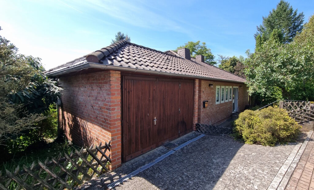 Das Bild zeigt eine Garage aus rotem Backstein an einem sonnigen Tag. Die Garage hat ein Satteldach mit dunklen Ziegeln und Dachrinnen. Vor der Garage befindet sich ein breites, braunes Holztor. Links neben dem Tor ist ein schmaler, niedriger Holzzaun sichtbar, der mit der Wand der Garage verbunden zu sein scheint. An der rechten Seite des Gebäudes befinden sich mehrere rechteckige Fenster mit weißen Rahmen. Davor wächst ein dichter, grüner Busch mit gelben Blüten. Weiter rechts ist ein weiterer Holzzaun mit einem Rautenmuster zu sehen. Der Boden vor der Garage ist mit grauem Pflaster bedeckt, auf dem sich ein langer, schmaler Metallschacht mit Lamellen befindet, der wohl der Entwässerung dient. Auf dem Dach der Garage sind zwei Schornsteine zu erkennen. Umgeben ist die Garage von üppiger Vegetation. Links und im Hintergrund wachsen verschiedene Bäume und Sträucher mit dunkelgrünen Blättern. Der Himmel ist strahlend blau mit wenigen weißen Wolkenstreifen.