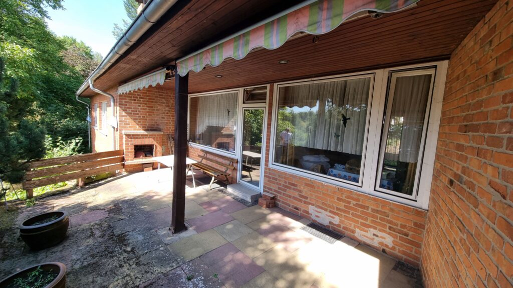 Die Aufnahme zeigt die Außenseite eines Hauses mit einer Ziegelwand, einem überdachten Bereich und einem Garten. Im Vordergrund sind zwei runde Blumentöpfe zu sehen, einer davon mit einer Pflanze gefüllt. Der Boden besteht aus unregelmäßigen Steinplatten in verschiedenen Rottönen und Gelbtönen, die teilweise von Schatten durchzogen sind. Links neben den Blumentöpfen steht eine einfache Holzbank. Der überdachte Bereich wird von einem Dach mit dunklen Holzverkleidungen und einer Markise mit vertikalen Streifen in Grün, Rosa und Hellblau getragen, die sich wellenförmig erstreckt. Eine dunkle Säule stützt das Dach. Dahinter befindet sich die Hauswand, die aus roten Ziegeln gemauert ist. An der Wand sind mehrere große Fenster mit weißen Rahmen angebracht, die mit hellen, durchscheinenden Vorhängen verdeckt sind. Durch die Fenster sind im Inneren Möbel und ein Tisch zu erkennen, und man sieht auch die Reflexion von Bäumen und Himmel. Im rechten Teil des Bildes sind zwei schwarze Lüftungsgitter im Boden eingelassen. Links neben dem überdachten Bereich ist eine Außenkaminattrappe aus Ziegelsteinen zu sehen, in deren Öffnung einige Holzscheite liegen. Eine weitere Holzbank steht links vom Kamin. Im Hintergrund erstreckt sich ein dichter, grüner Baum- und Buschbestand unter einem hellblauen Himmel. Die Sonne scheint und wirft helle Schatten auf den Boden und die Ziegelwand.