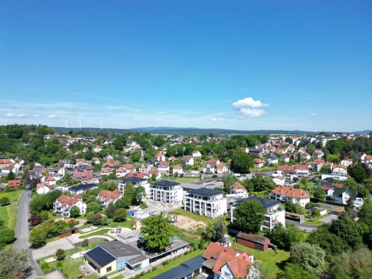 Auf dieser Drohnenaufnahme ist ein Stadtteil von Schwalmstadt-Treysa zu sehen. Der Himmel ist strahlend blau mit kleinen Wölkchen im Hintergrund. Ebenfalls im Hintergrund links sind drei Windräder zu erkennen. Auf der Aufnahme sind viele Wohnhäuser mit überwiegend roter Dacheindeckung zu sehen. Der Stadtteil ist sehr grün mit vielen Großbäumen. Vereinzelt sind Straßen und Wege zu erkennen. In der Bildmitte sind vier moderne Mehrfamilienhäuser mit dunkler Dacheindeckung zu sehen. Die Wohnhäuser sind nach Süden ausgerichtet und verfügen über großzügige Balkone. Im Dachgeschoss erkennt man sehr große teilweise überdachte Dachterrassen. Im Vordergrund ist ein großes Grundstück mit Spielgeräten und einem größeren Flachbau zu erkennen. Hier handelt es sich um einen Kindergarten.